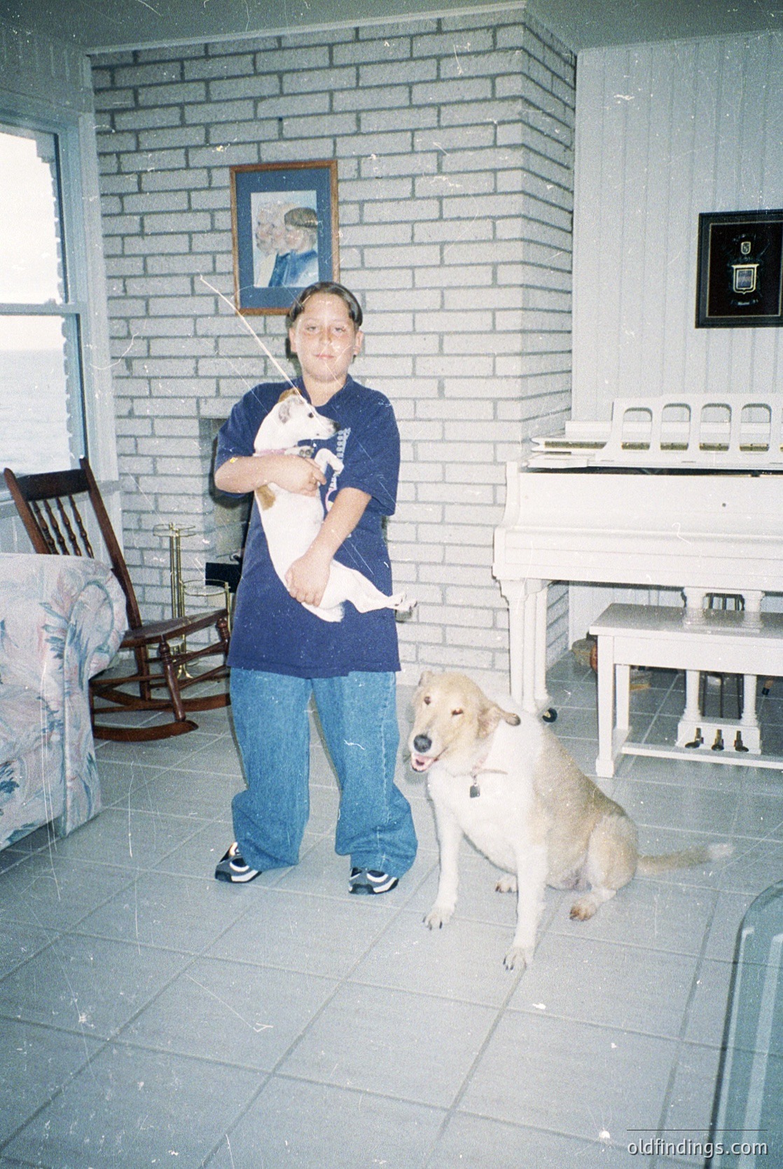 Mid-20th century indoor scene: Woman in a dark blue T-shirt and jeans holds a white cat, standing beside a light-colored dog on tiled flooring. Brick-paneled wall with a framed portrait above a vintage piano and wooden chair. Warm, lived-in domestic atmosphere.