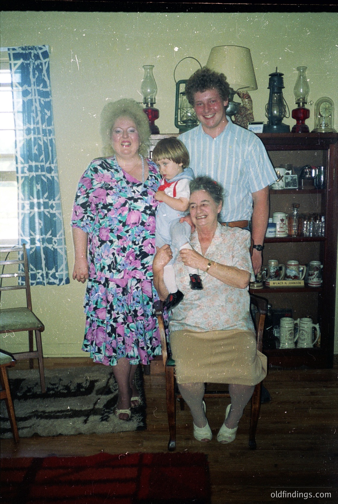 Family portrait in a mid-century kitchen, featuring four generations: a grandmother in a floral dress, seated; a woman in a patterned dress holding a toddler; a man in a striped shirt; and a baby in a white outfit. Decor includes vintage lamps, ceramic mugs, and a patterned curtain. Likely 1960s–1970s, USA.