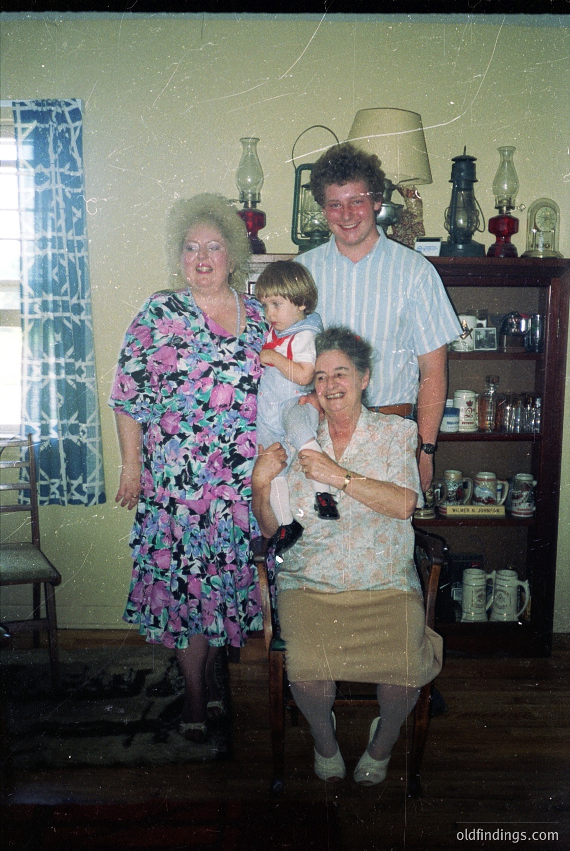 Family portrait in a mid-century kitchen, featuring three adults and a child. The woman on the left wears a floral-patterned dress, while the seated woman in the foreground has a vintage wristwatch and a patterned blouse. The man stands behind them in a striped shirt. Decor includes vintage lanterns, ceramic mugs, and a patterned curtain. Likely 1960s–1970s, Western household.