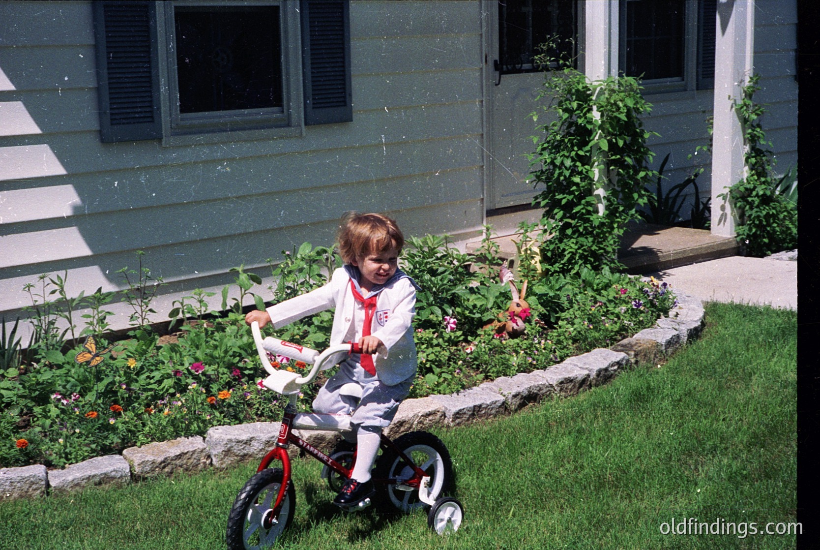 Young child in 1970s-style training wheels outfit rides red bicycle on lush suburban lawn. White vinyl siding house with closed shutters and blooming flowerbeds in foreground. Classic mid-century American backyard scene.
