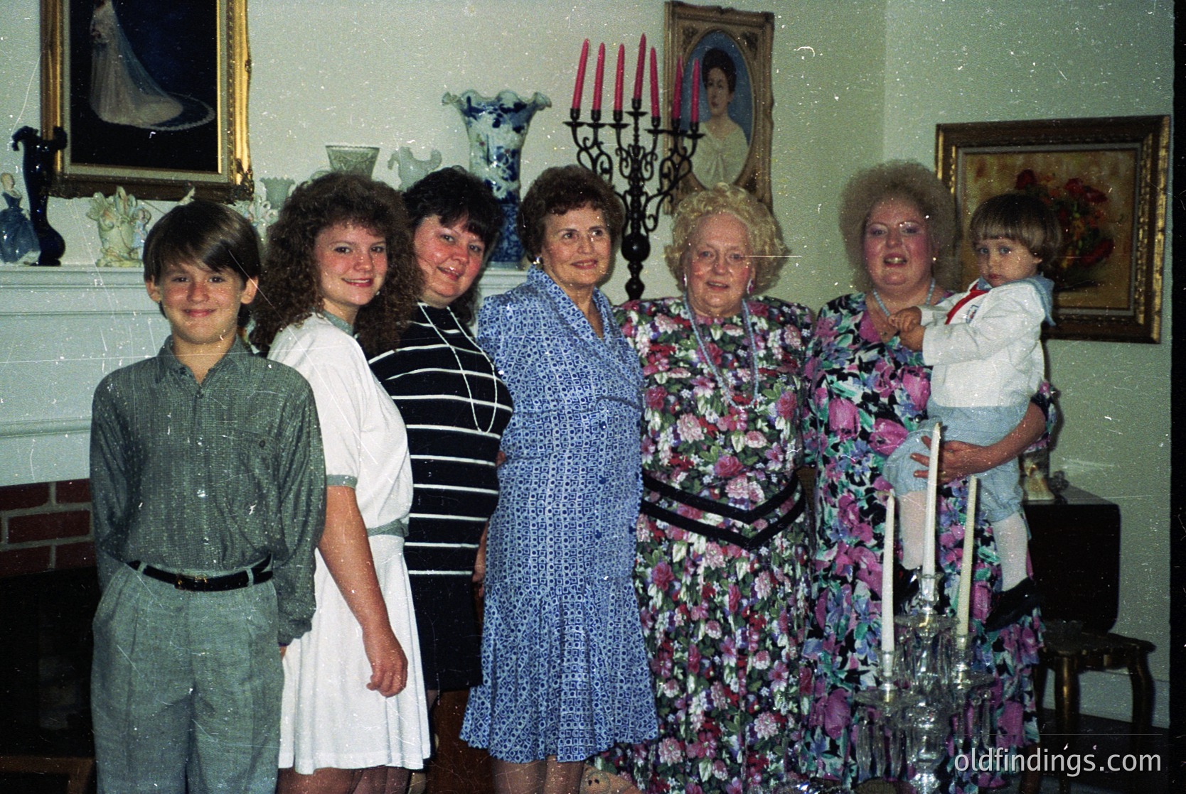 Family portrait from the 1980s–1990s, featuring five adults and two children in a dimly lit indoor setting. The group stands in front of a fireplace with framed religious artwork, including a portrait of a saint. Clothing styles suggest mid-late 20th century: floral dresses, striped blouses, and a child’s white dress. Candelabra and vintage decor enhance nostalgic atmosphere.