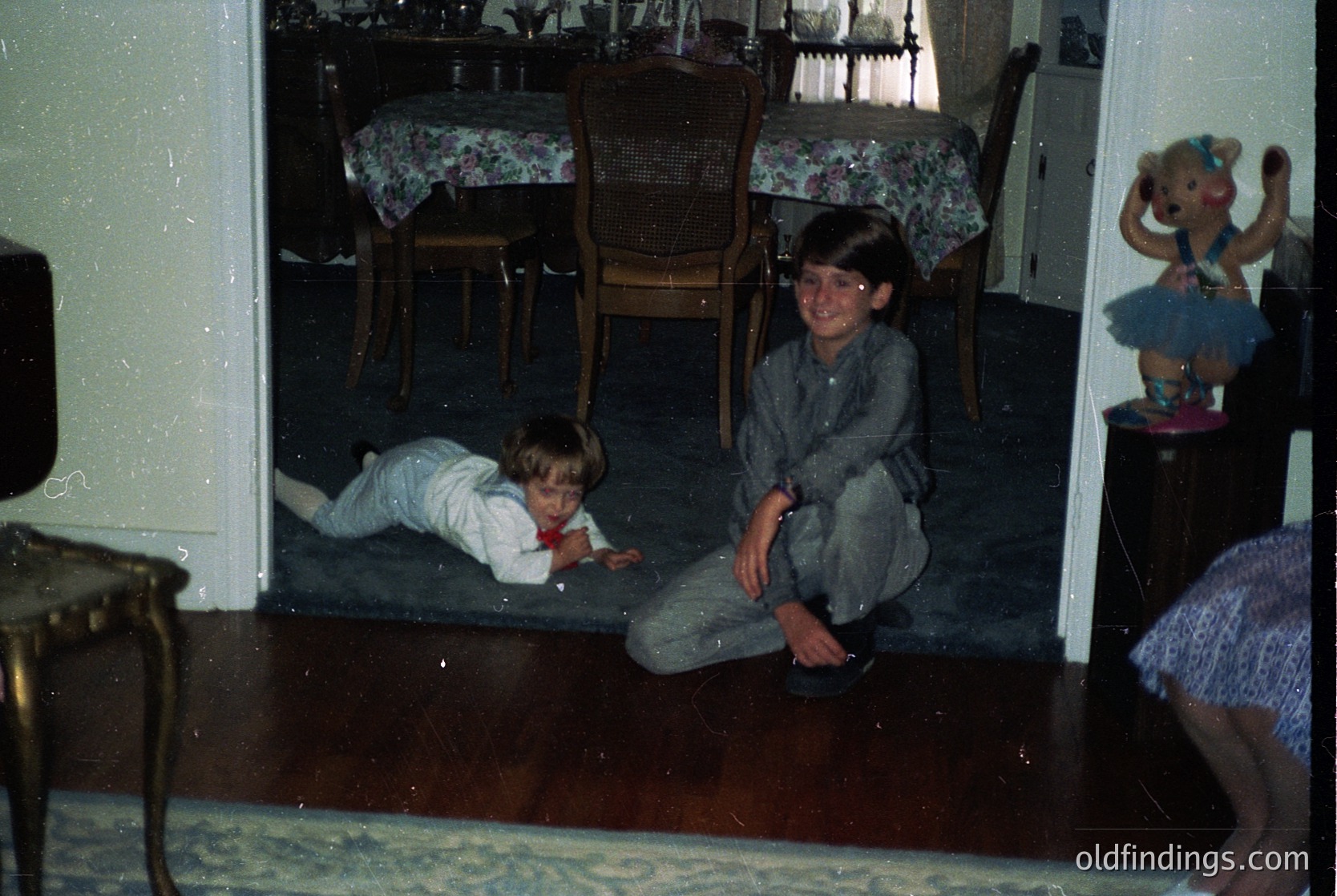 Vintage indoor scene featuring two children playing on a wooden floor in a mid-century dining area. The boy sits cross-legged in overalls, while the toddler crawls on a patterned rug. A floral tablecloth, cane-backed chairs, and a stuffed animal on a shelf suggest a family home from the **1970s**. Warm lighting and classic decor evoke nostalgia.