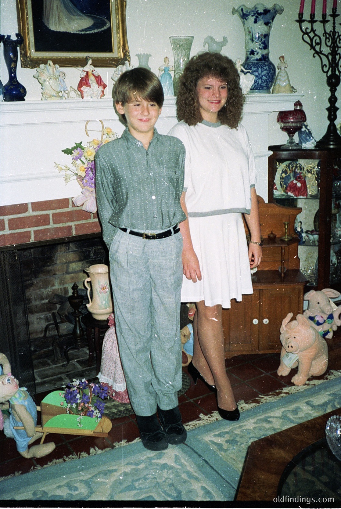 Two children pose indoors in a vintage-style living room, likely 1970s–1980s. Boy wears a button-down shirt, pleated trousers, and loafers; girl in a white knee-length dress with a belt. Surroundings include a fireplace with decorative urns, floral arrangements, and a fireplace mantel adorned with porcelain figurines. A wooden cabinet with glass doors displays ceramic items. Rug features floral patterns, and a plush teddy bear sits on the floor. écor