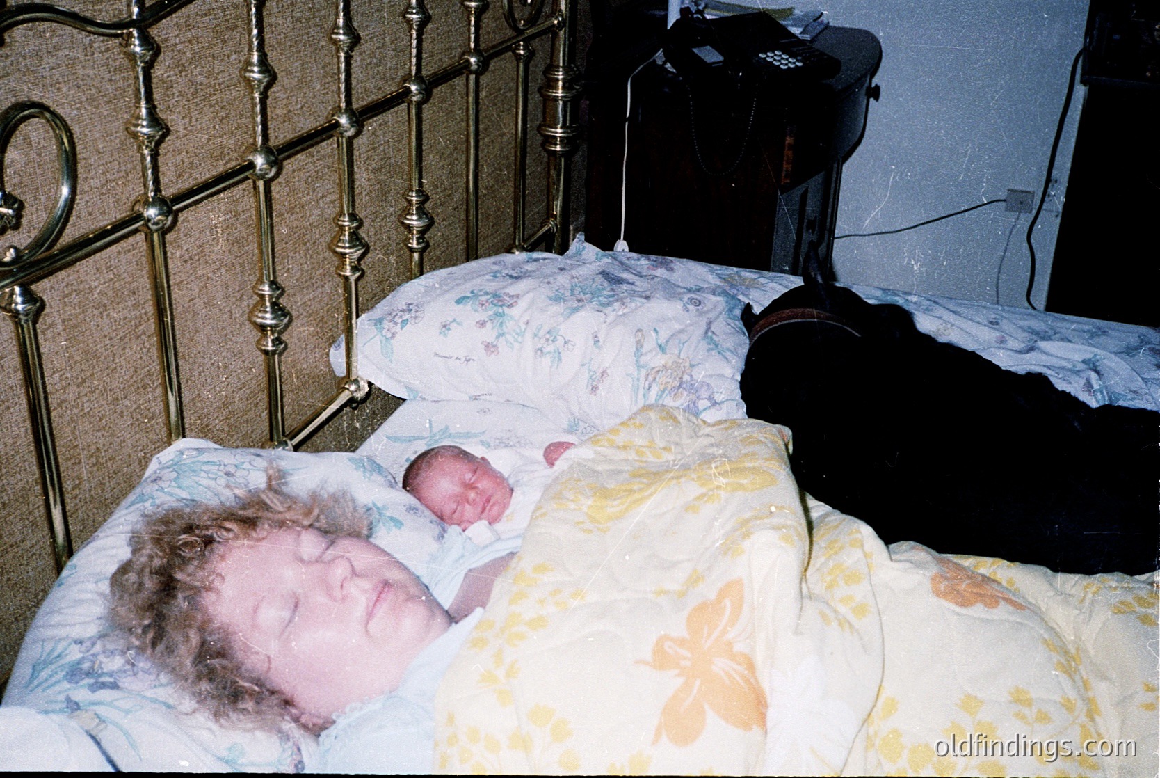 Vintage hospital bed scene featuring a newborn swaddled in floral bedding, alongside a sleeping adult. Ornate metal bed frame with curved posts suggests mid-20th century medical design. Dark suitcase and medical equipment hint at institutional setting.