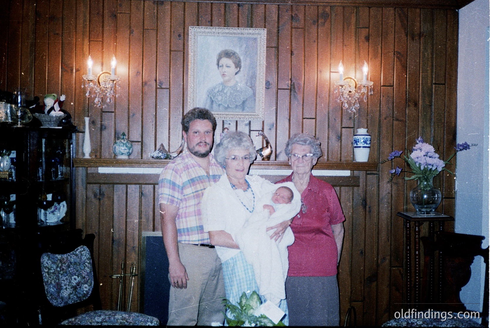 Family portrait in a rustic wooden-paneled interior, likely 1980s–1990s. Three adults pose with a newborn, framed by vintage wall sconces, floral arrangements, and a framed portrait above. Warm lighting enhances the homely, intimate atmosphere.
