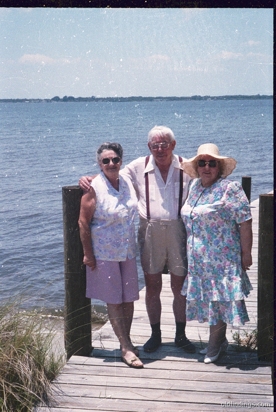 Three seniors pose on a wooden pier by a calm sea, mid-20th century. Man in light-colored trousers and suspenders stands between two women—one in a floral dress with a straw hat, the other in a patterned blouse and purple skirt. Sunlight and clear skies enhance the serene seaside atmosphere.