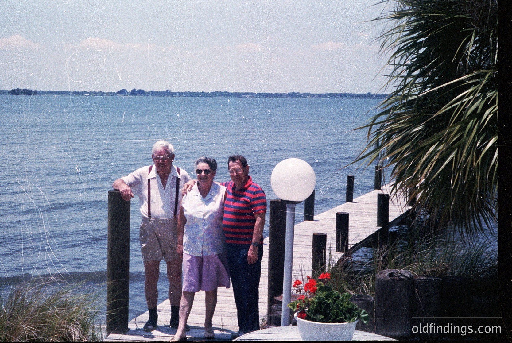 Three adults pose on a seaside wooden pier, 1970s-80s. Elderly couple in retro swimwear (man in suspenders, woman in floral dress) flank a man in striped shirt. Palm trees and potted flowers frame the scene.