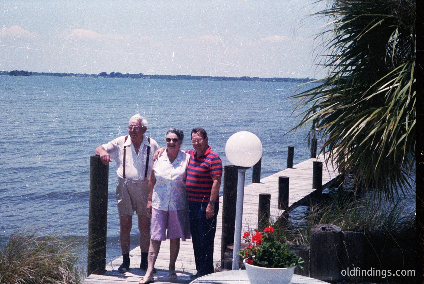 Three adults pose on a seaside wooden pier, 1970s–1980s coastal USA. Man in striped shirt, woman in floral dress, and man in light jacket stand beside a white lamp post. Clear blue water and distant land visible. Pier bordered by potted plants and palm fronds.
