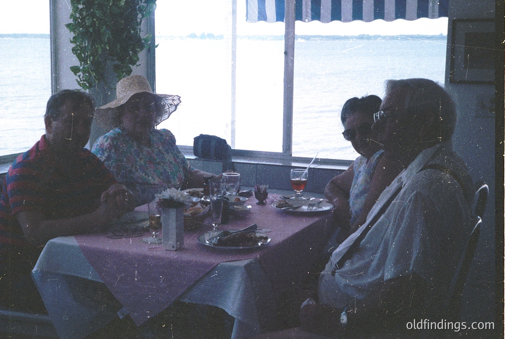 Four adults seated at a seaside restaurant table, enjoying a meal with drinks. Mid-century dining ambiance with red tablecloths, framed mirrors, and a view of the ocean through large windows. Glasses, plates, and cutlery suggest a leisurely meal. Likely or coastal dining.