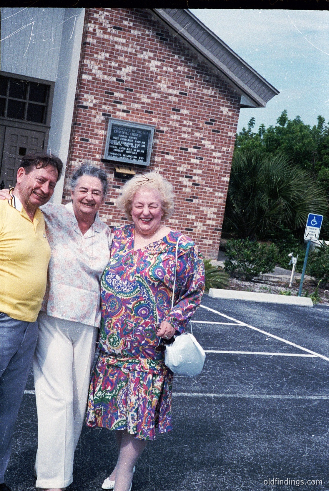 Three elderly individuals pose outside a brick building with a plaque, likely from the 1970s. The woman in the center wears a floral-patterned dress, while the man on the left sports a yellow shirt and white pants. The setting appears suburban, with palm trees and a handicap parking sign visible.
