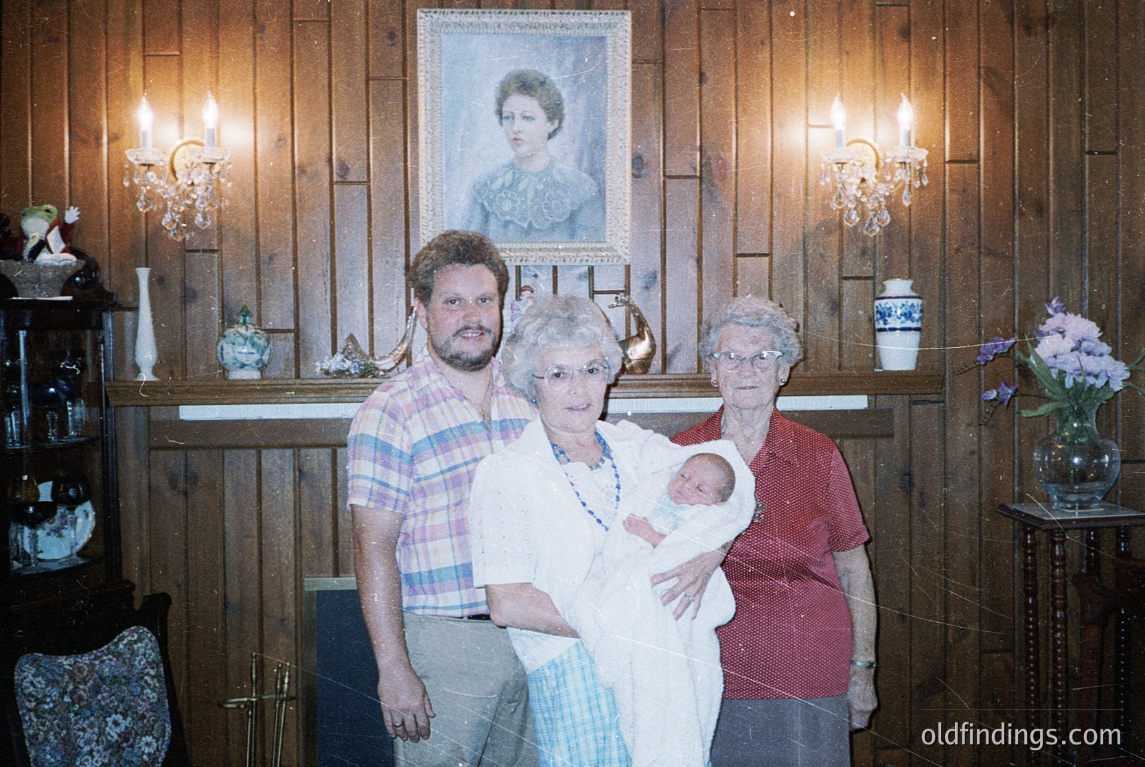Family portrait in a rustic wooden interior, likely 1980s–1990s. Three adults and a newborn stand in front of a framed portrait of a woman, surrounded by vintage chandeliers and floral vases. Warm lighting enhances the nostalgic, intimate atmosphere.