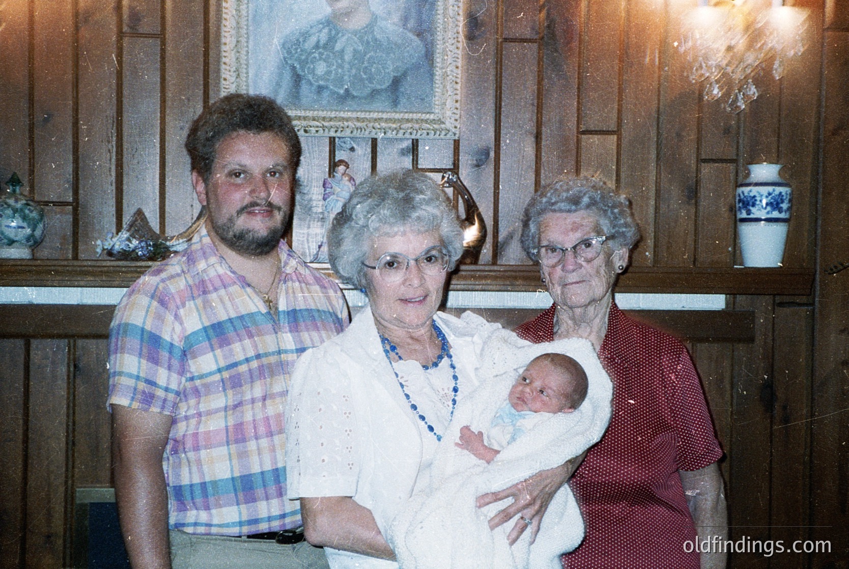 Family portrait featuring a man in a plaid shirt, two elderly women in formal attire (one holding a grandchild), and a framed painting in the background. Likely late 20th century (1980s-1990s), indoor setting with warm lighting.