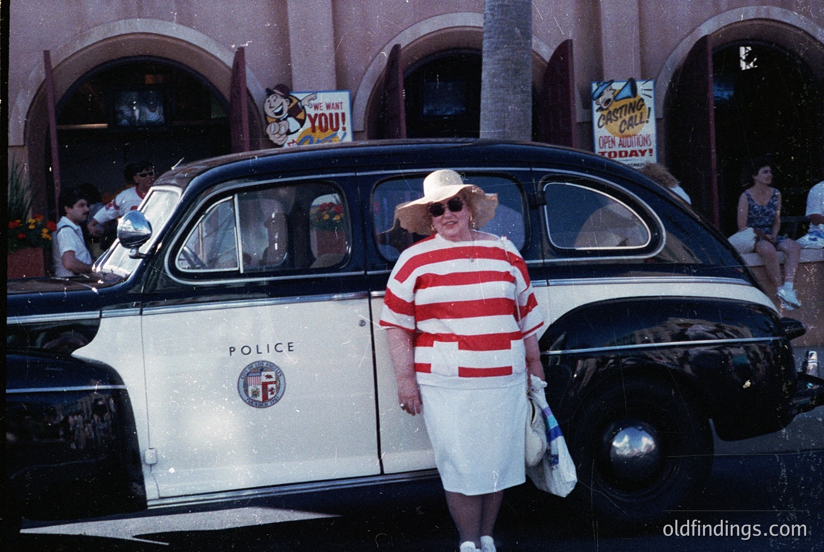 Vintage 1950s-60s police car with "POLICE" insignia parked near a retro-themed arcade. Woman in red-and-white striped dress and sun hat poses beside the vehicle. Brightly lit signage advertises "You Bet You!" and "Cocktail Lounge." Crowd in summer attire visible in background.