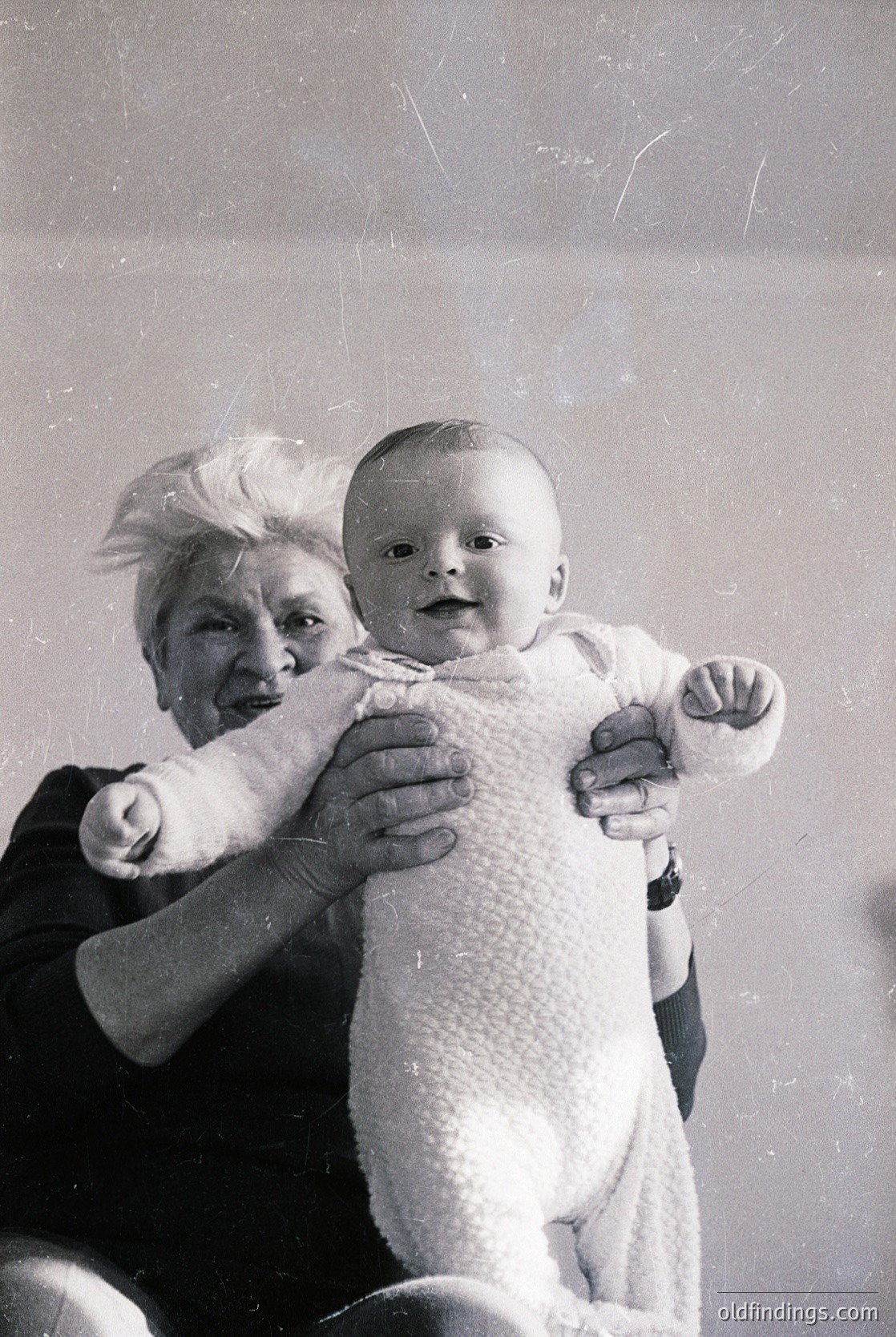 Vintage black-and-white portrait of an elderly woman holding an infant, both smiling directly at the camera. The woman’s hairstyle and clothing suggest a mid-20th-century setting, likely 1950s–1960s. The baby wears a knitted onesie with a textured pattern.