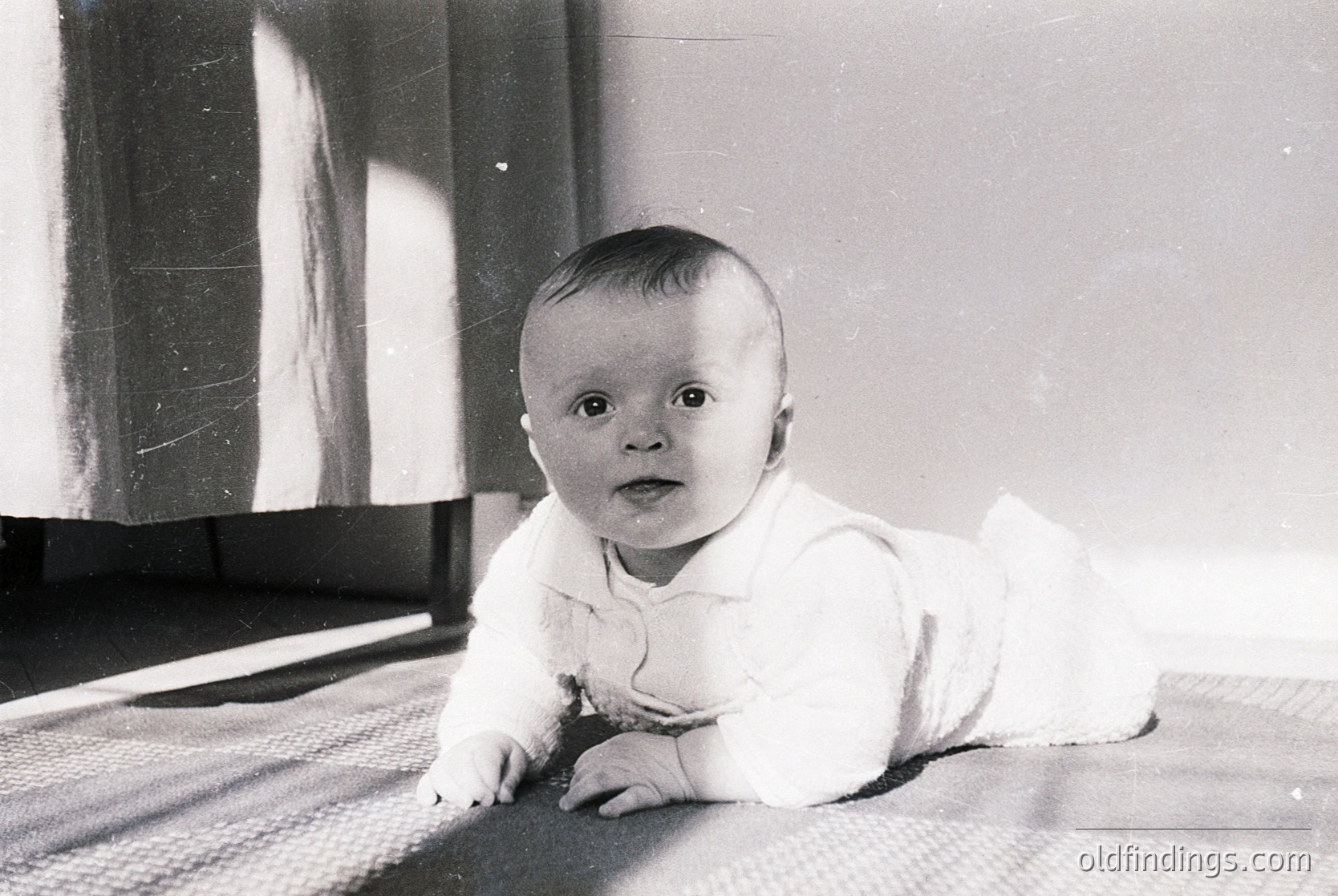 Vintage black-and-white portrait of an infant crawling on a patterned rug, dressed in a white onesie with a bow. Soft indoor lighting highlights delicate facial features. Likely mid-20th century (1940s–1960s) based on style and grain.