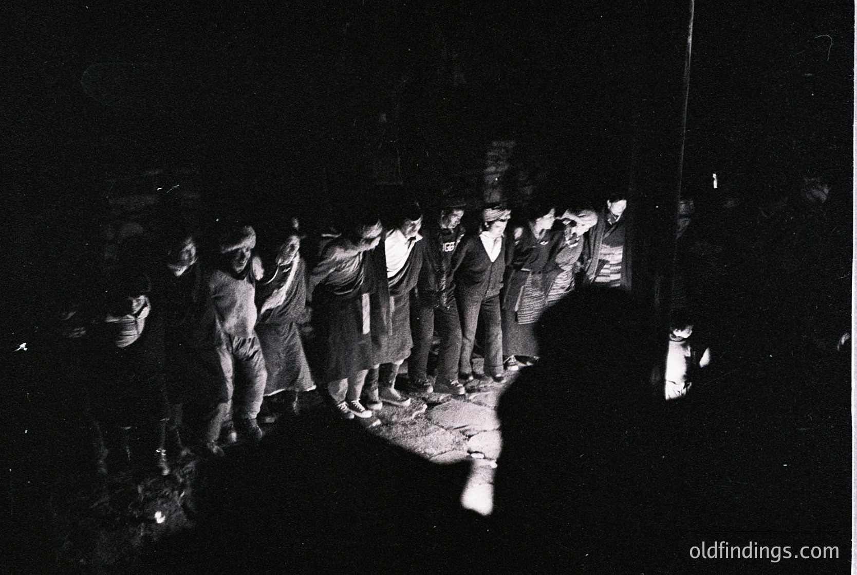 Black-and-white street scene capturing a group of men in formal attire (suits, ties, hats) standing in a line under dim lighting, possibly a 1950s-1960s urban setting. Shadows and low contrast emphasize the gathering’s solemnity. Potential historical or cultural event context.