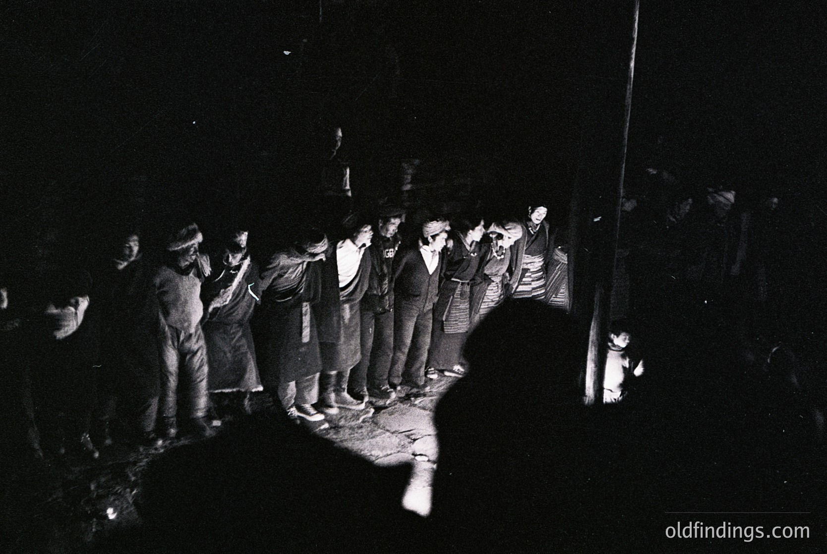 Black-and-white photo of a nighttime protest or demonstration, likely 1960s–1980s. Crowd of men in dark clothing, some holding signs with text (partially legible). Urban setting with visible streetlights and shadows. Atmosphere suggests civil unrest or political gathering.