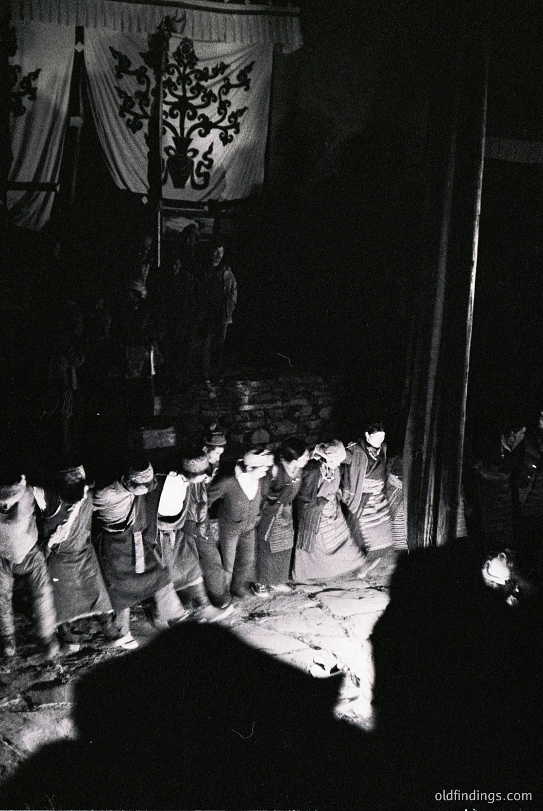 Black-and-white nighttime scene capturing a traditional procession with a decorated float featuring an ornate floral emblem. Crowd in vintage attire, likely 1950s–1970s, gathered around a central figure holding a torch. Urban setting with visible architecture and streetlights. Evokes cultural heritage and communal celebration.