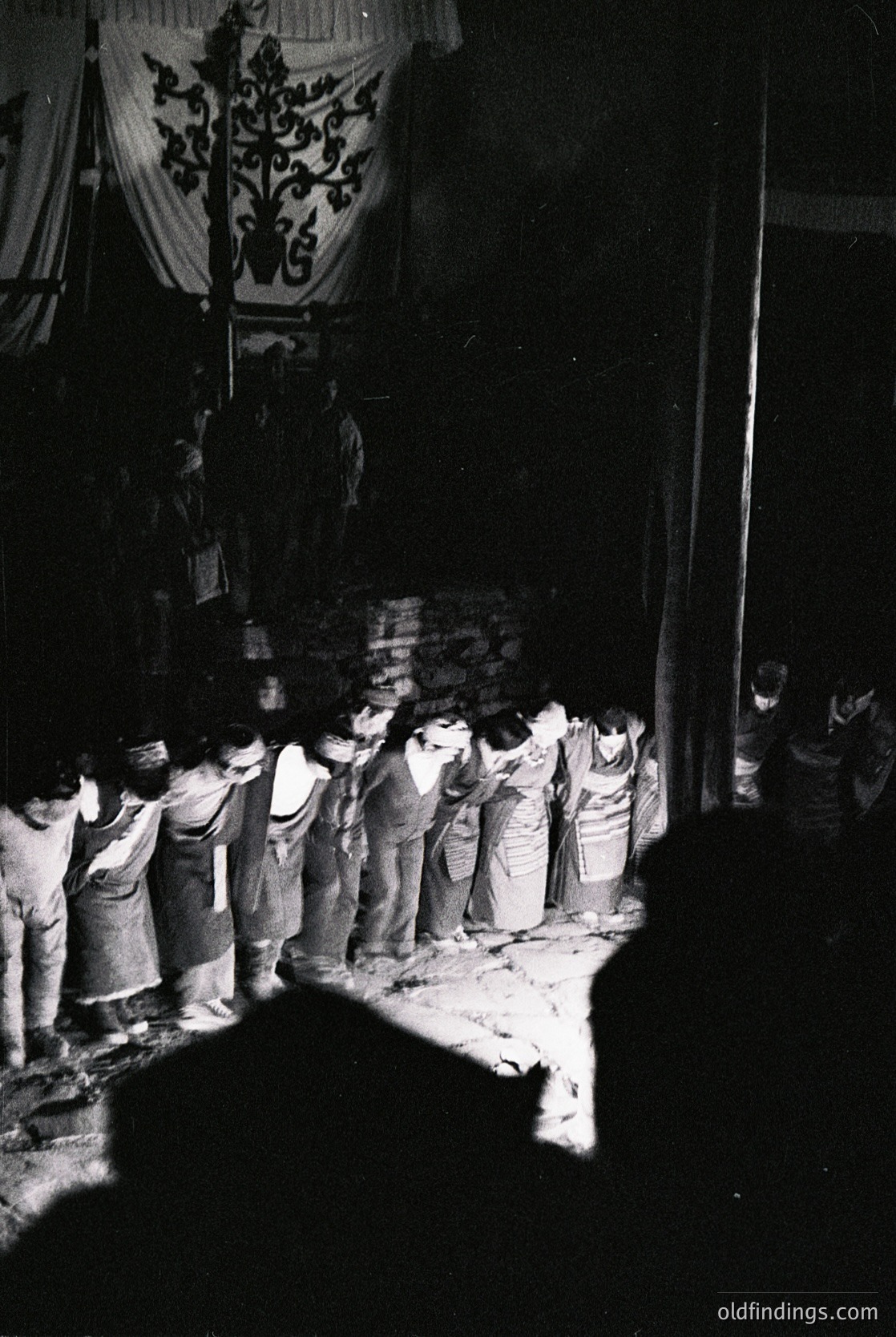 Black-and-white photograph of a solemn procession in a dimly lit courtyard. A group of men in light, loose-fitting clothing and head coverings kneel or bow toward a central figure draped in a patterned cloth banner. The scene suggests a ritual or ceremonial gathering, likely Eastern European or Balkan in origin, circa mid-20th century.