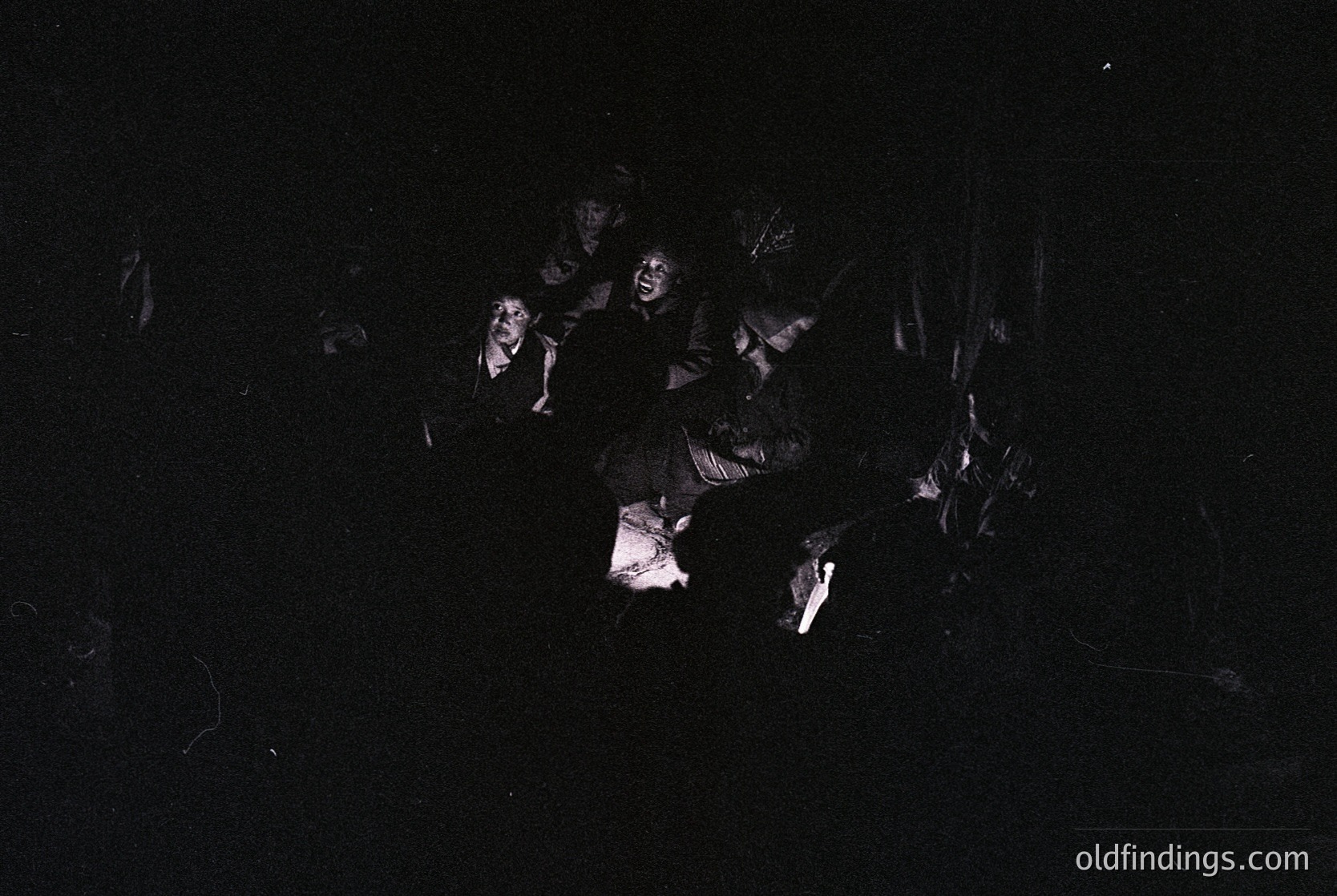 High-contrast black-and-white image of a group of people in a dimly lit, possibly indoor setting. Silhouetted figures appear engaged in a seated or kneeling arrangement, with one central figure illuminated by a faint light source. Style suggests documentary or candid photography.