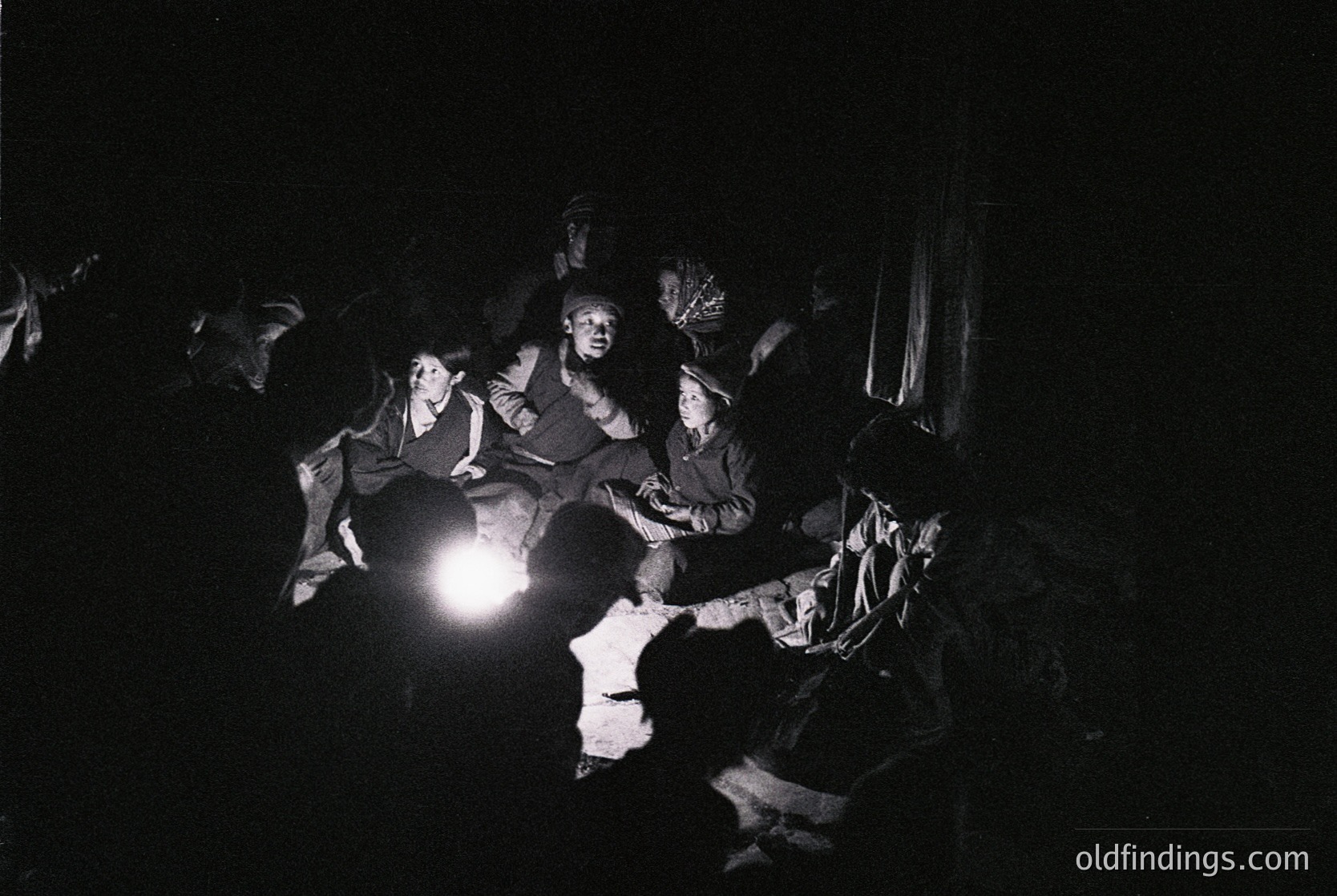 Black-and-white nighttime gathering in a dimly lit, enclosed space. Six individuals seated on wooden benches, illuminated by a single handheld flashlight. Central figure in a dark coat and hat directs attention, surrounded by others in winter attire—scarves, coats, and hats. Likely a rural or communal setting, possibly a folk or traditional event. Style suggests 1950s–1970s Eastern European or Soviet-era photography.