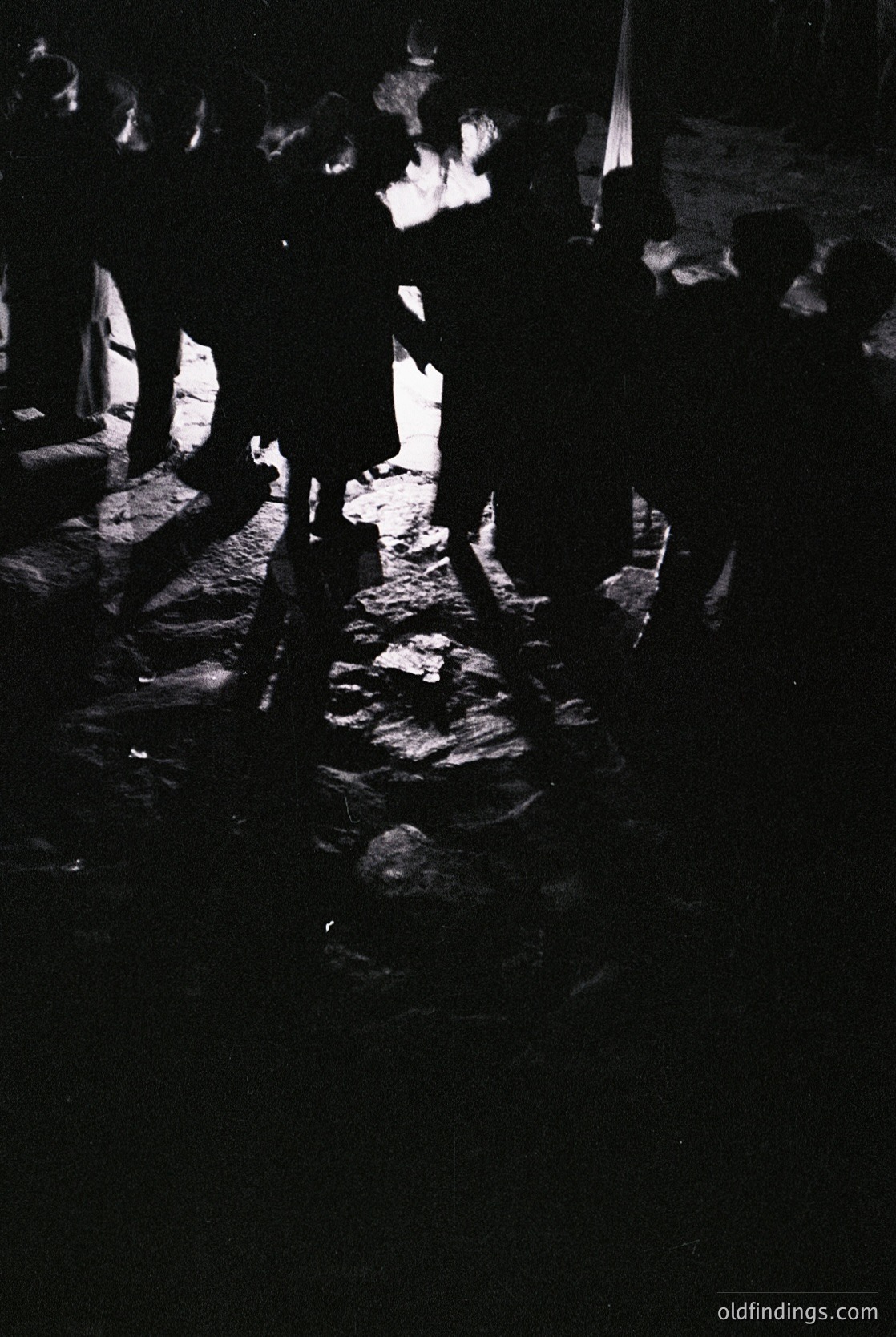 Silhouetted crowd dancing around a central fire pit on cobblestone ground, likely a traditional folk festival. Low lighting enhances dramatic contrast.