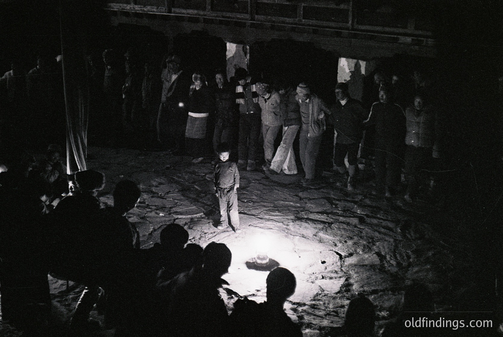 Black-and-white nighttime gathering in an open-air setting, likely a mine or industrial site. Central figure in white shirt and pants stands illuminated by a single light source, surrounded by a crowd in dark clothing. Formal attire suggests a ceremonial or commemorative event. Mid-20th century (1950s–1970s) Eastern European industrial heritage.