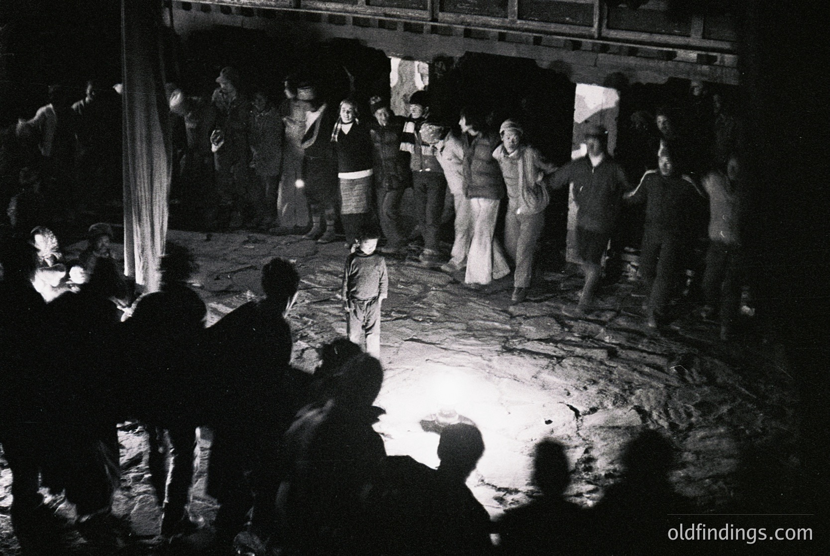 Black-and-white photo captures a lively 1970s dance scene in an indoor venue, likely a club or community hall. Crowd of ~30 people, mostly young adults in flared pants, bell-bottoms, and patterned tops, dancing under dim lighting. Central figures in white tops and dark pants appear to lead a group dance. Concrete floor and exposed beams suggest utilitarian architecture.