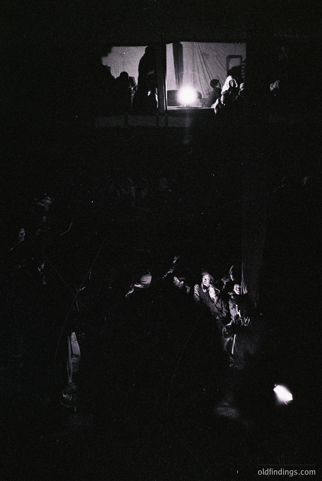 Low-angle shot of a dimly lit indoor crowd, silhouetted against a bright stage light. Central figures appear to be performing or speaking, with one person’s raised arms visible. Likely a concert, lecture, or theatrical performance in a confined space. Style suggests 1970s–1980s documentary or concert photography.