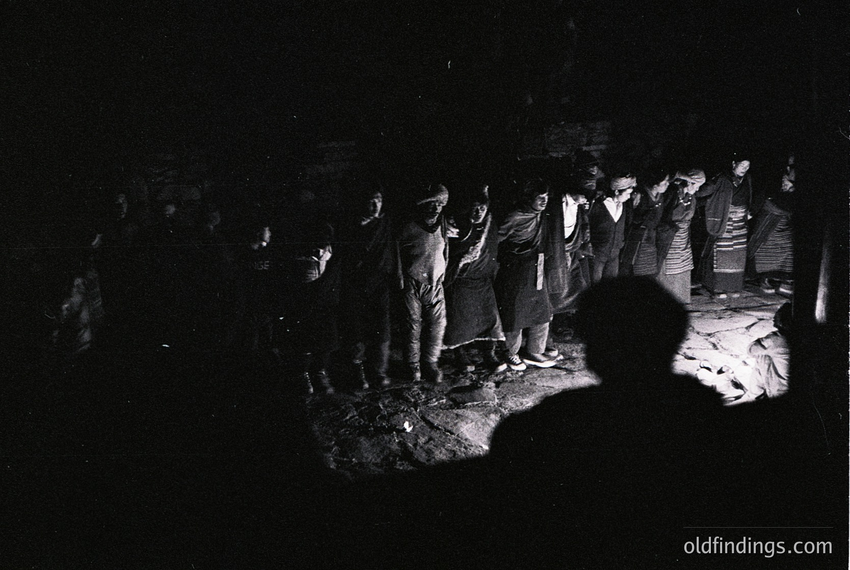 Black-and-white nighttime gathering in a dimly lit, enclosed space with a central fire. Group of men in traditional attire, possibly ceremonial or ritualistic, standing in a semi-circle. One figure kneels near the fire, suggesting a communal or cultural event. Likely Eastern European or Balkan region, mid-20th century.