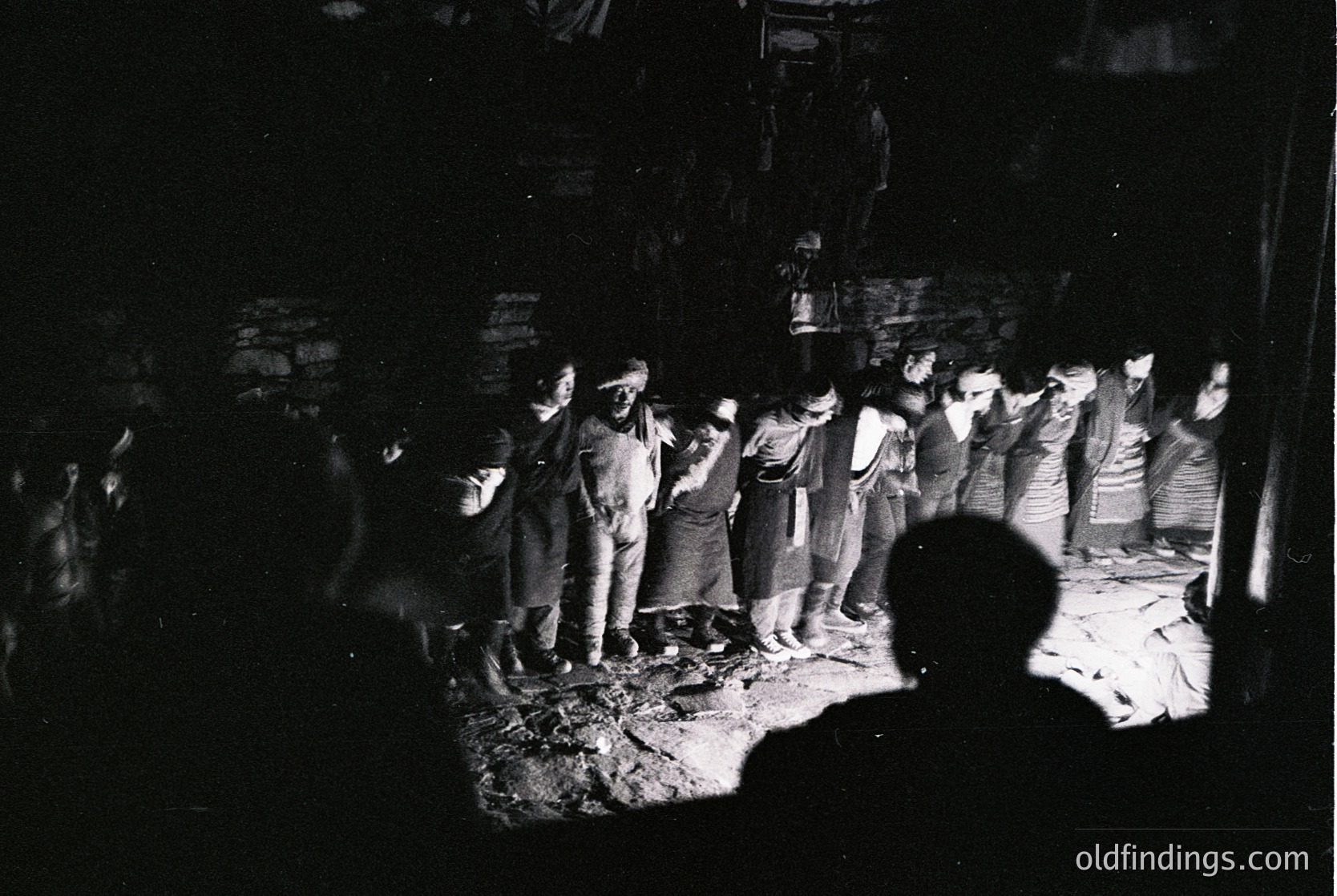 Black-and-white street scene featuring a crowd of men in 1960s–70s attire, likely Eastern European. Central figures wear suits and headscarves, standing near a low wall or rubble. Shadows and low lighting suggest nighttime or poor visibility. Urban setting with visible signage in Cyrillic script.
