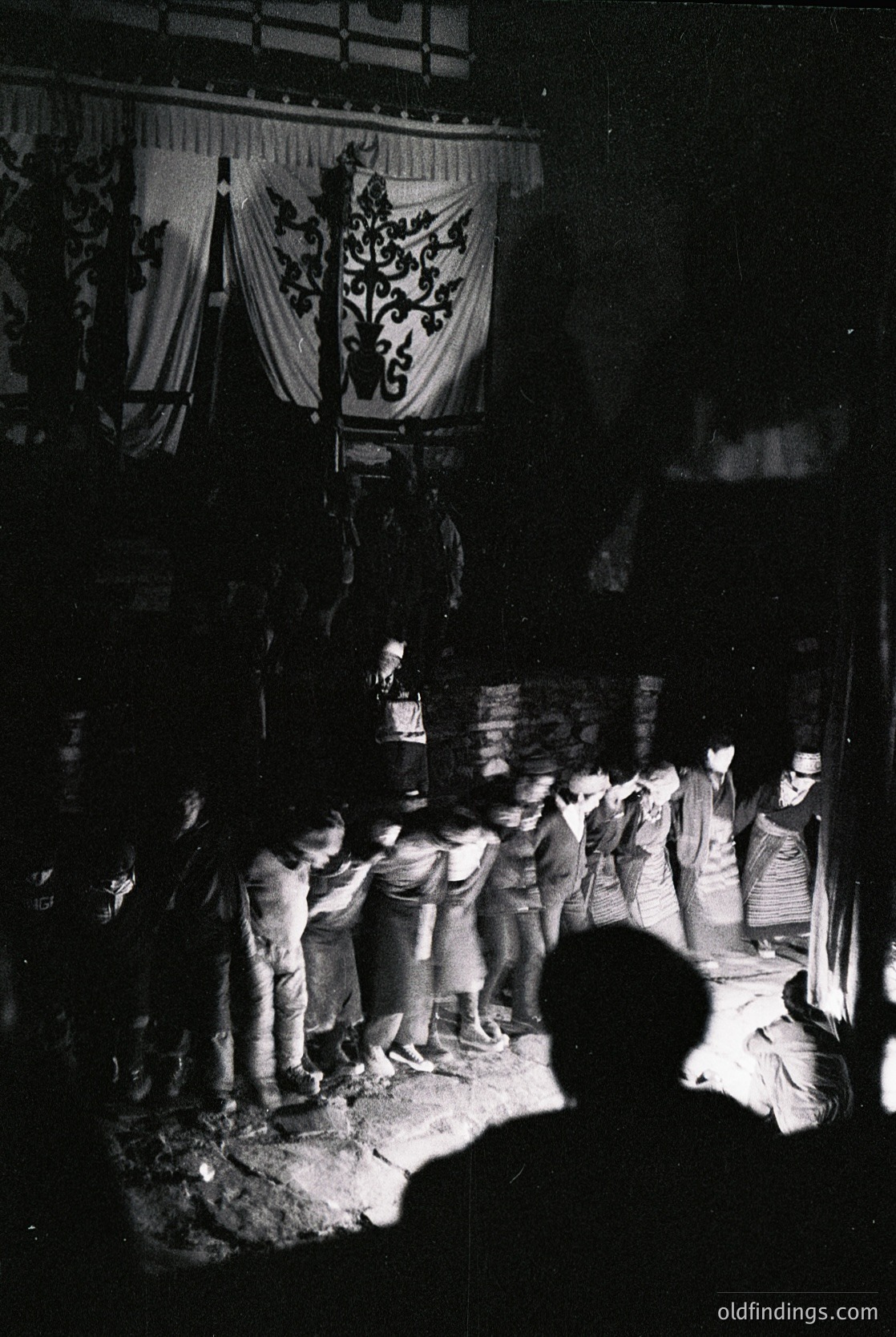 Black-and-white nighttime gathering under illuminated banner featuring a heraldic emblem (crossed swords, crown, and "1275"). Crowd of ~20 people in 1960s-70s attire (bell-bottoms, jackets, hats) gathered around a central figure holding a bottle. Wooden structure and lanterns suggest a festive or ceremonial event.