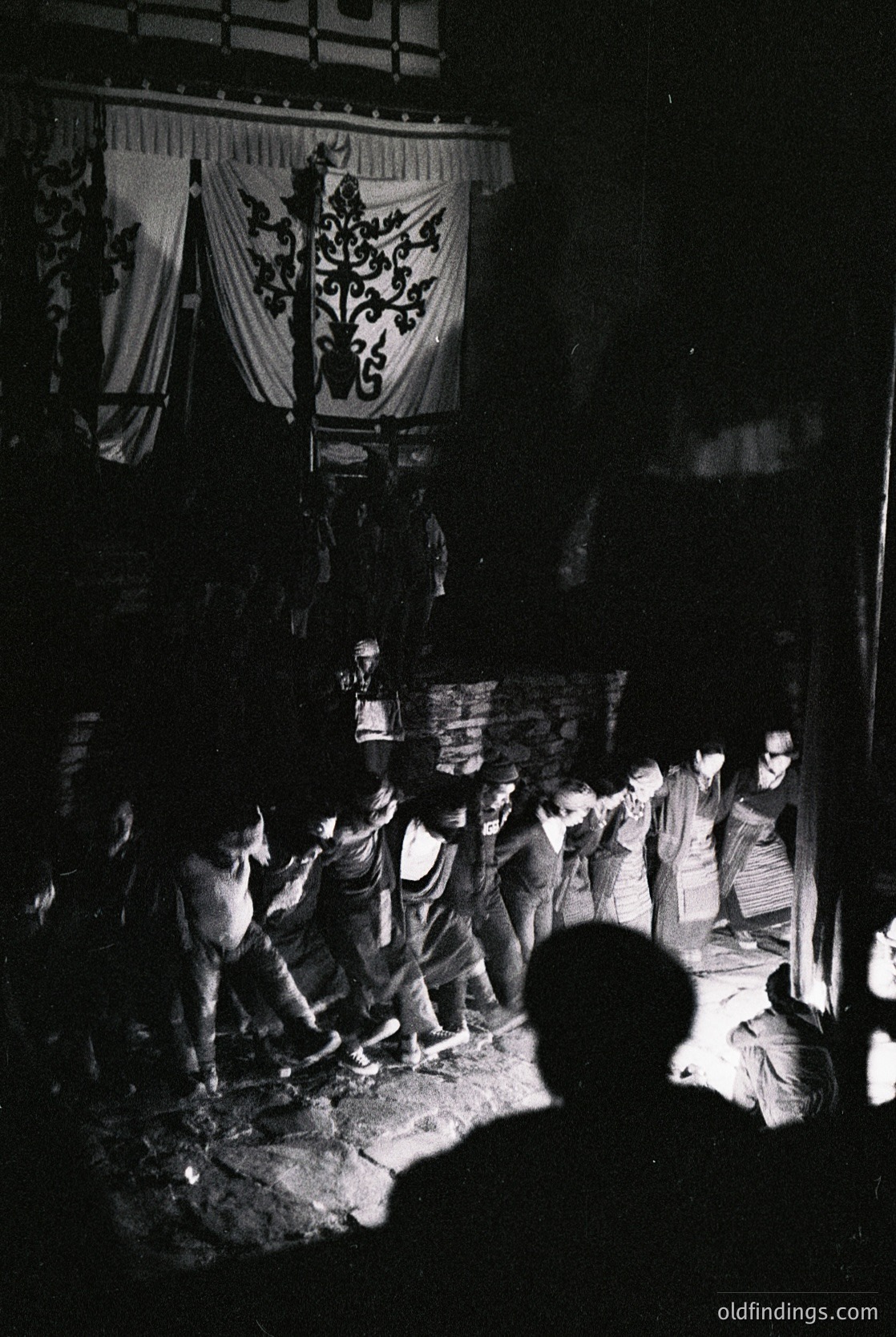 Black-and-white nighttime scene of a traditional Japanese festival procession. Participants carry a large, ornate banner with a floral crest and Japanese characters. Crowd gathers around a wooden structure, possibly a shrine or stage, under dim lantern light. Costumes suggest .
