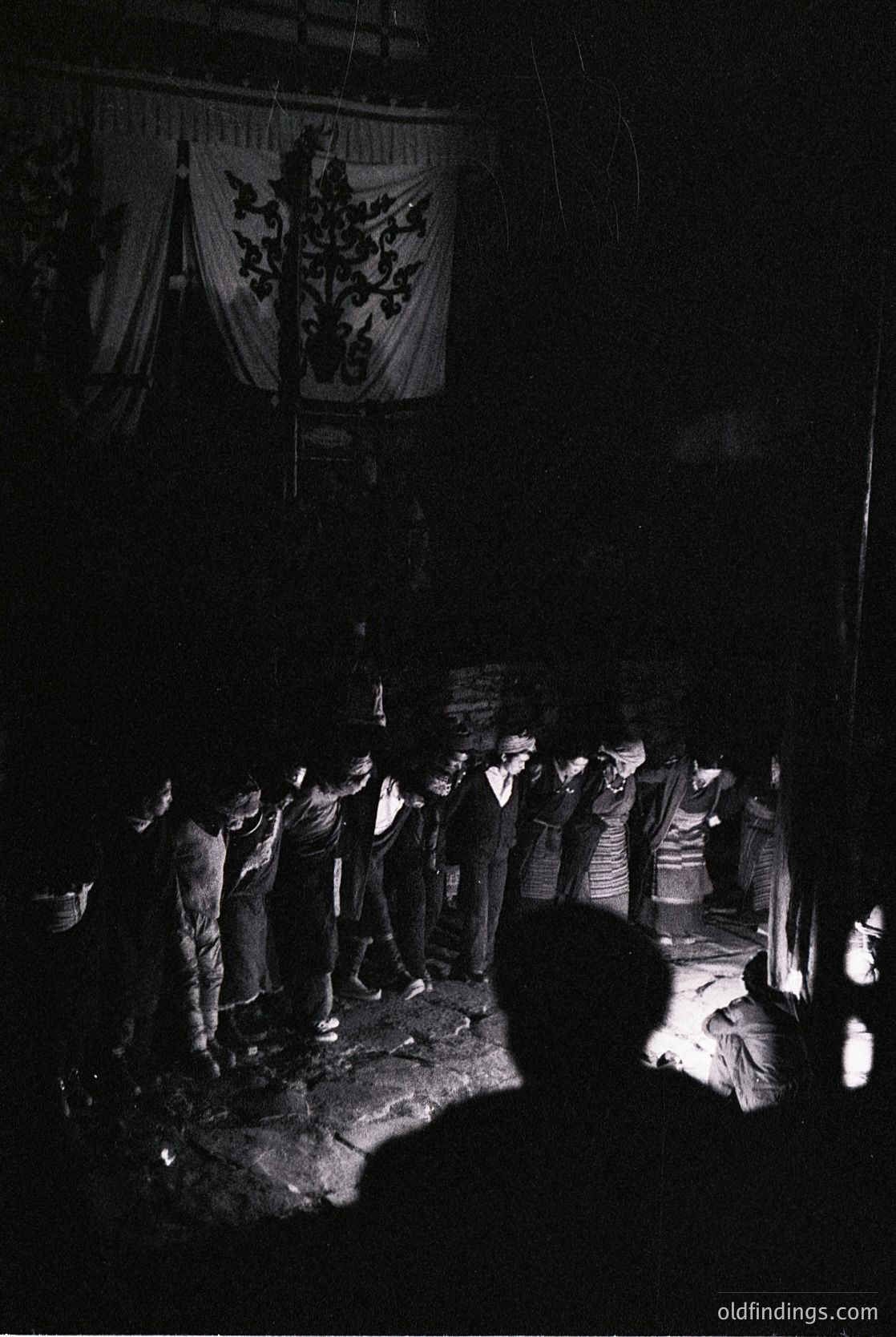 Group gathers around a central fire in a dimly lit courtyard, illuminated by flames. Embroidered banner with floral/foliage motif hangs overhead. Men in 1960s-70s attire (vests, ties, trousers) stand in a semi-circle. Traditional gathering suggests folk or ritual event.