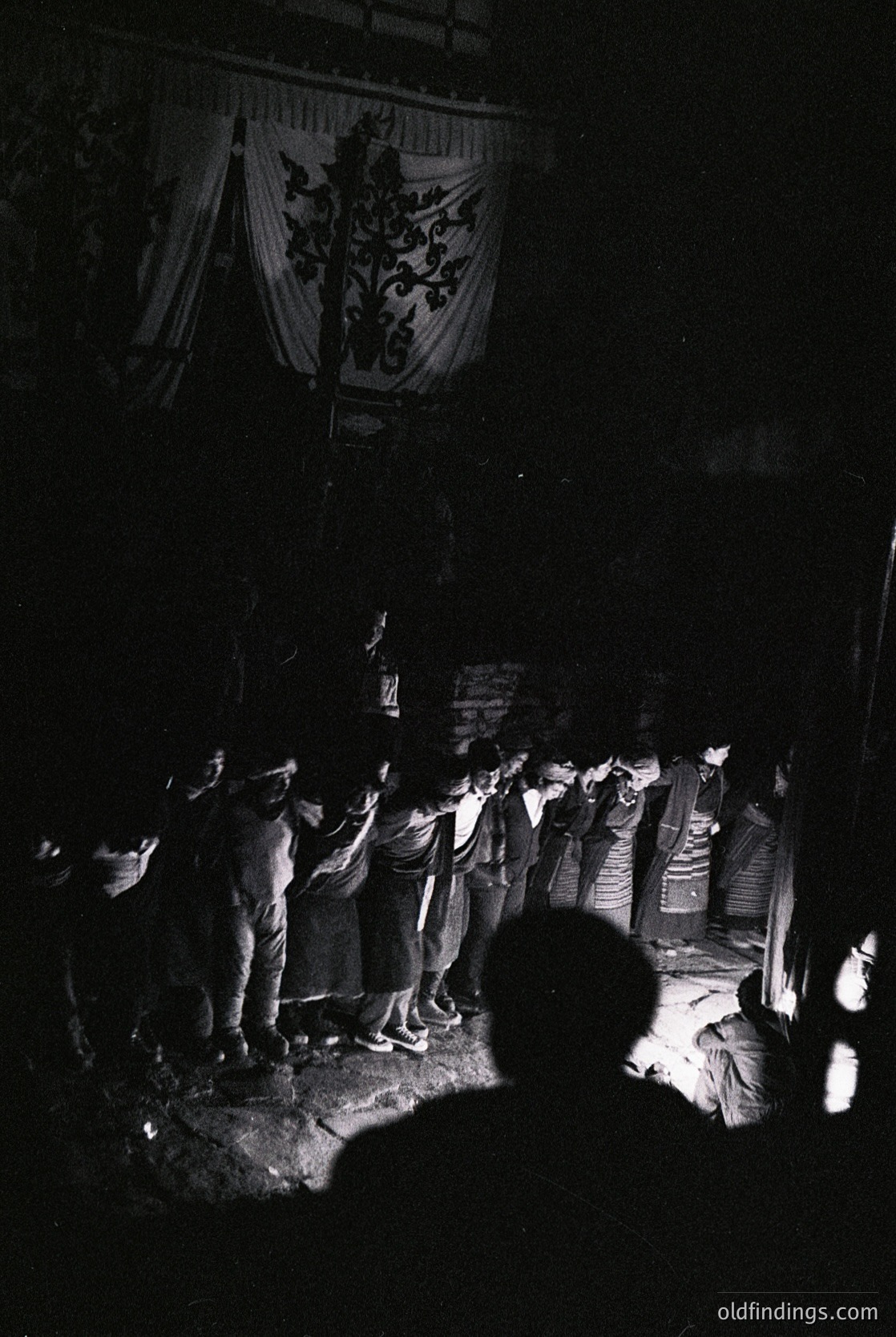 Black-and-white nighttime gathering under a banner featuring a crowned coat of arms, likely European heraldry. Crowd of men in 1960s–70s attire (short-sleeve shirts, trousers) forming a circle around a central figure holding a sign. Dim lighting from candles or lanterns illuminates faces and hands. Possible folk festival or traditional ceremony.