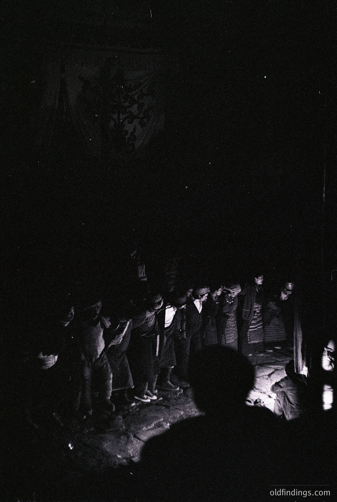 Black-and-white nighttime gathering in a forest clearing, featuring a semi-circle of seated and standing figures. Light source from a central fire illuminates faces and attire, suggesting a communal ritual or storytelling session. Mid-20th century outdoor cultural event, likely European.