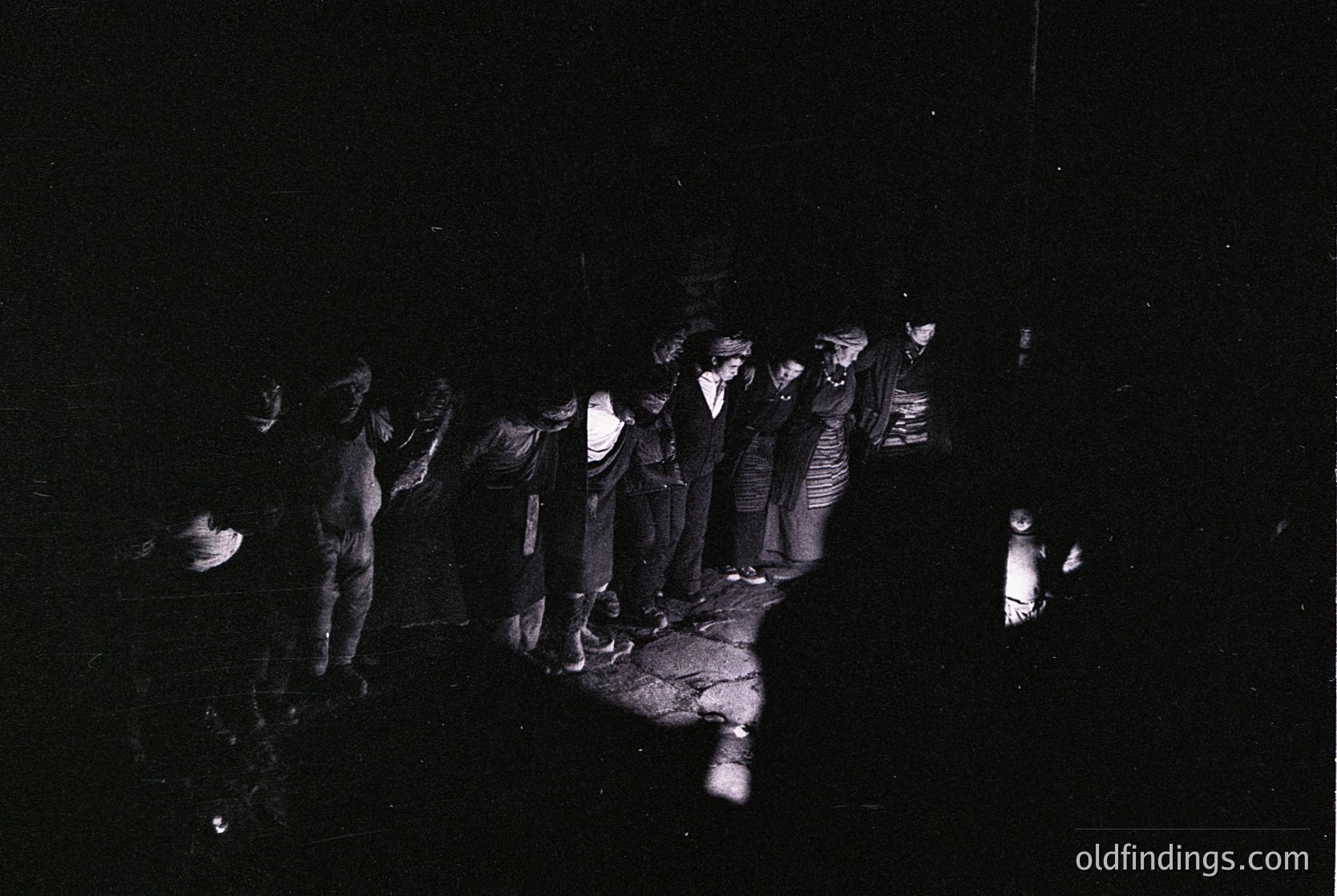 Black-and-white nighttime gathering of 12+ individuals in a dimly lit outdoor setting, illuminated by a single lantern. Group appears to be engaged in a communal activity, possibly a ritual or event. Mid-20th century attire suggests or . Rural or semi-rural location implied by surroundings.