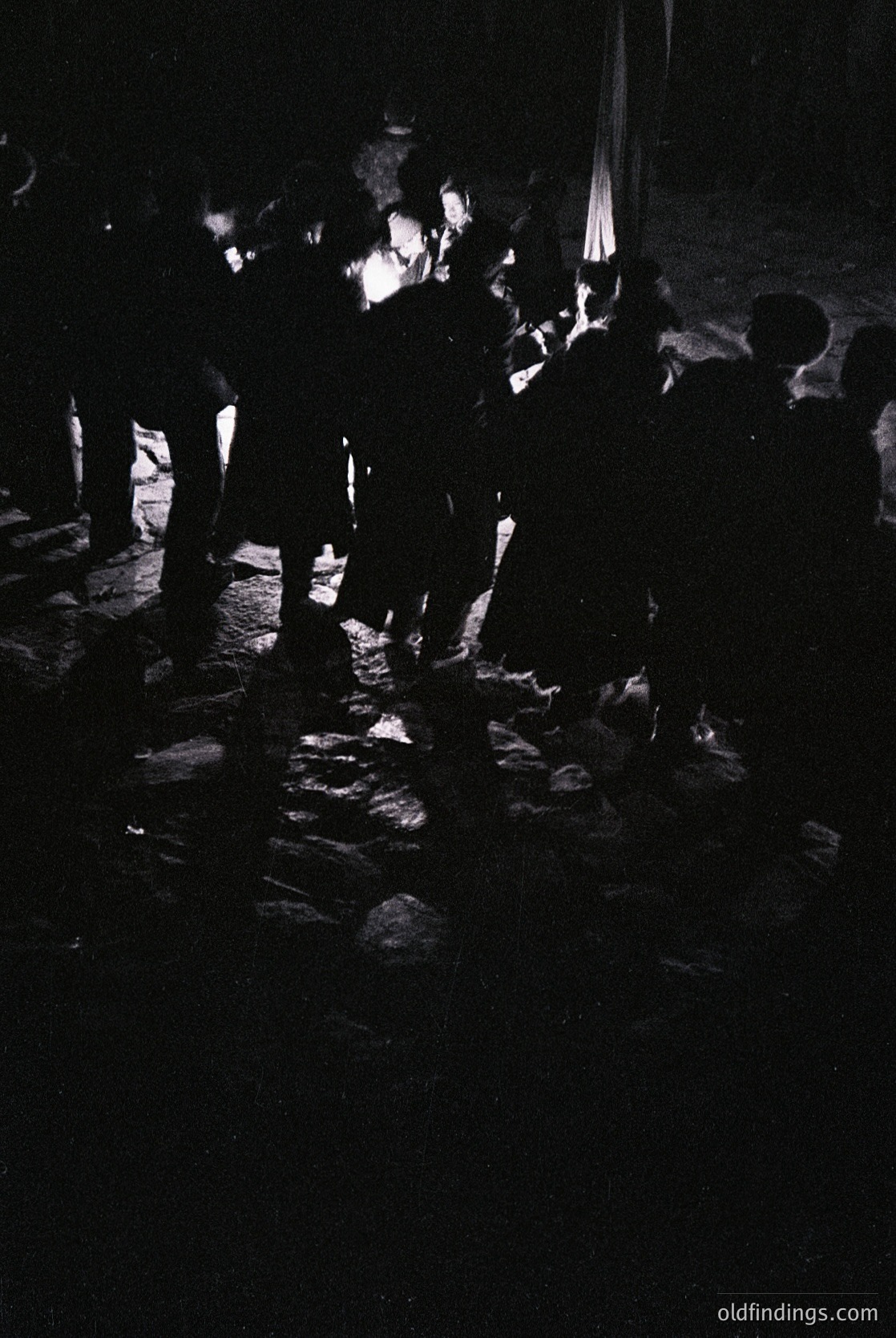 Silhouetted crowd gathered around a central fire in a dimly lit, cobblestone plaza. Silhouettes suggest evening or nighttime gathering, likely cultural/traditional event. Style hints at mid-20th century European setting.