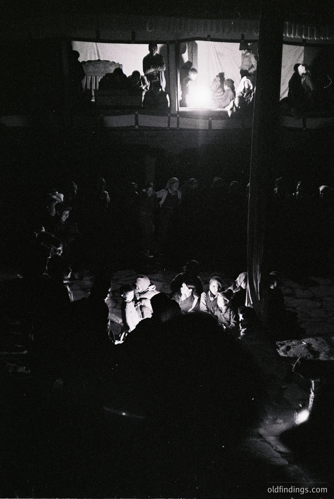 Vintage black-and-white photo of a dimly lit, two-tiered gathering with silhouetted figures. Lower level: small group huddled around a central light source, likely a lantern or candle. Upper level: larger crowd seated on wooden benches, facing inward. Architectural details include exposed wooden beams and a rustic interior. Evokes mid-20th century communal or festive atmosphere.