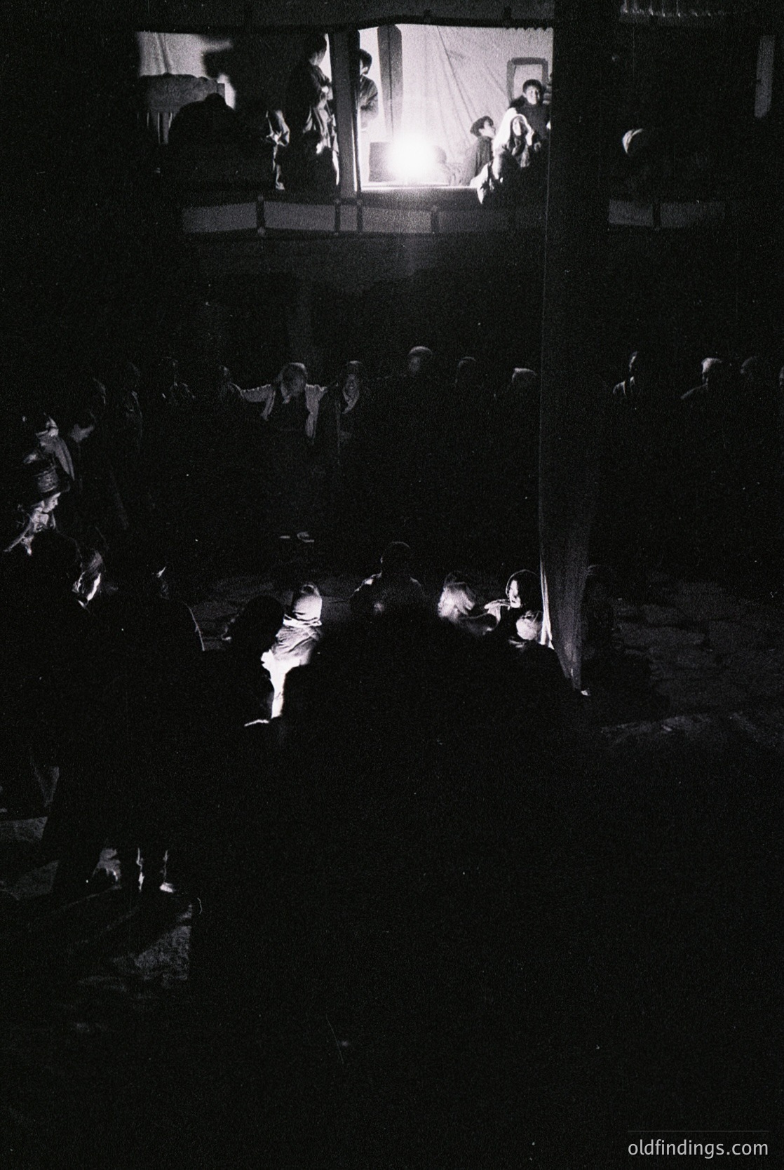 Vintage black-and-white shot of a dimly lit indoor gathering, likely a 1950s–1960s social event. Crowded room with silhouetted figures—some seated, others standing—facing a stage or elevated platform. Warm light source illuminates the upper area, suggesting a performance or speech. Casual attire hints at a communal or festive occasion.