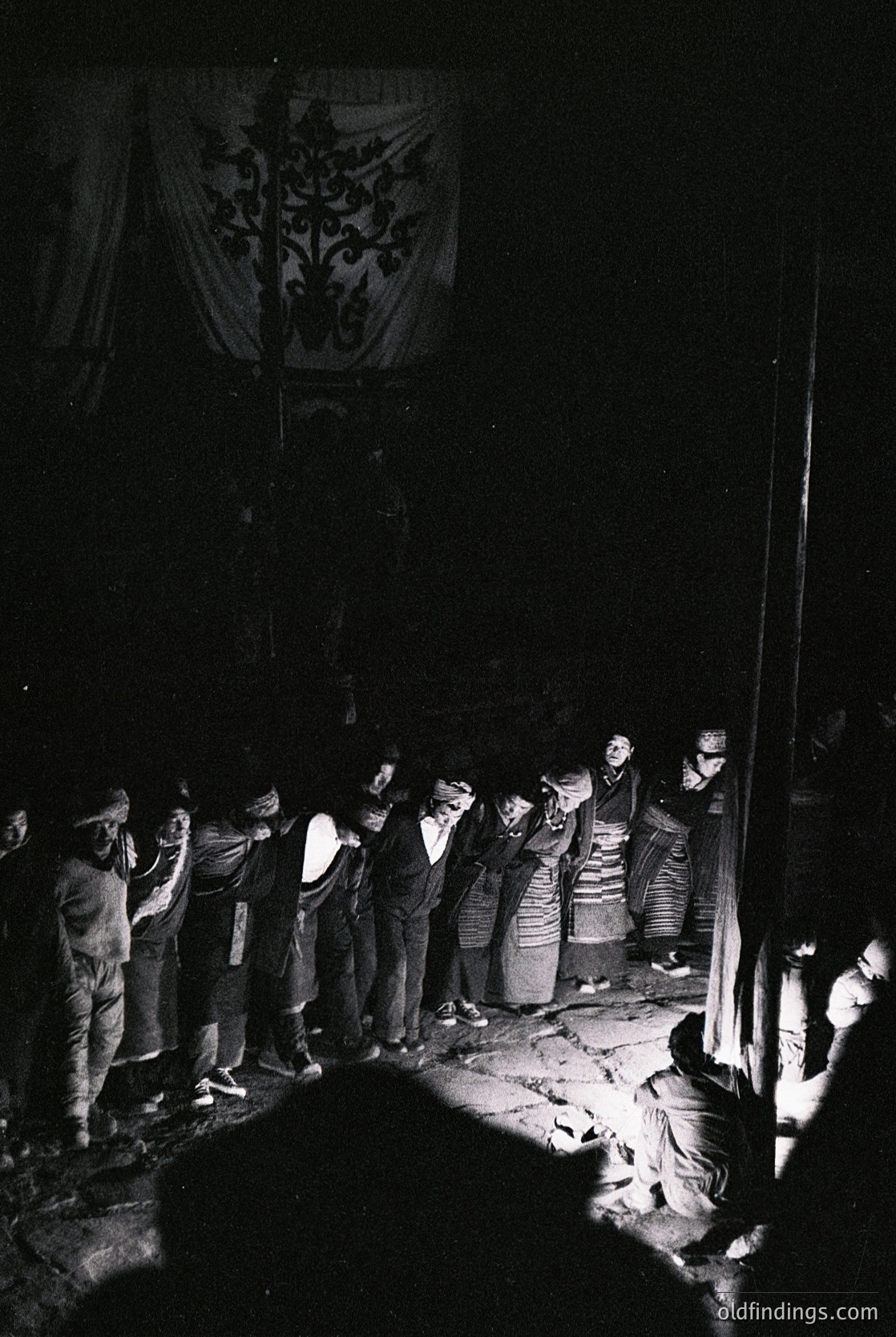 Black-and-white nighttime gathering under ornate embroidered banner, likely Bulgarian folk festival. Men and women in traditional attire—men in vests, women in layered skirts—form a circle around a central figure. Handheld lanterns illuminate faces, suggesting communal ritual or celebration. Mid-20th century (1950s–1970s) rural Bulgaria.