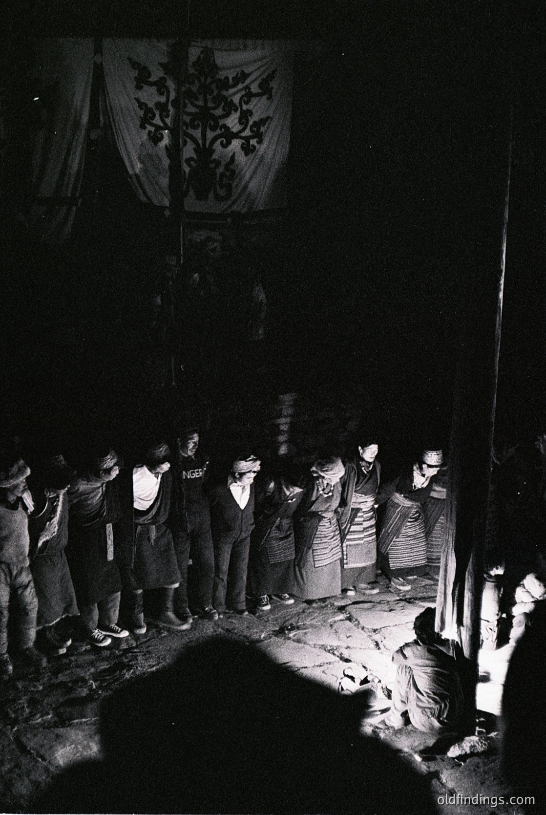 Indoor gathering featuring traditional attire, likely a folk ritual or ceremony. Men in striped shirts, women in layered skirts and headscarves. Central figure kneeling near a firelight source, surrounded by standing participants. Embroidered banner with floral motif hangs overhead. Mid-20th century Eastern European setting, possibly Bulgaria or Romania. Authentic cultural documentation.