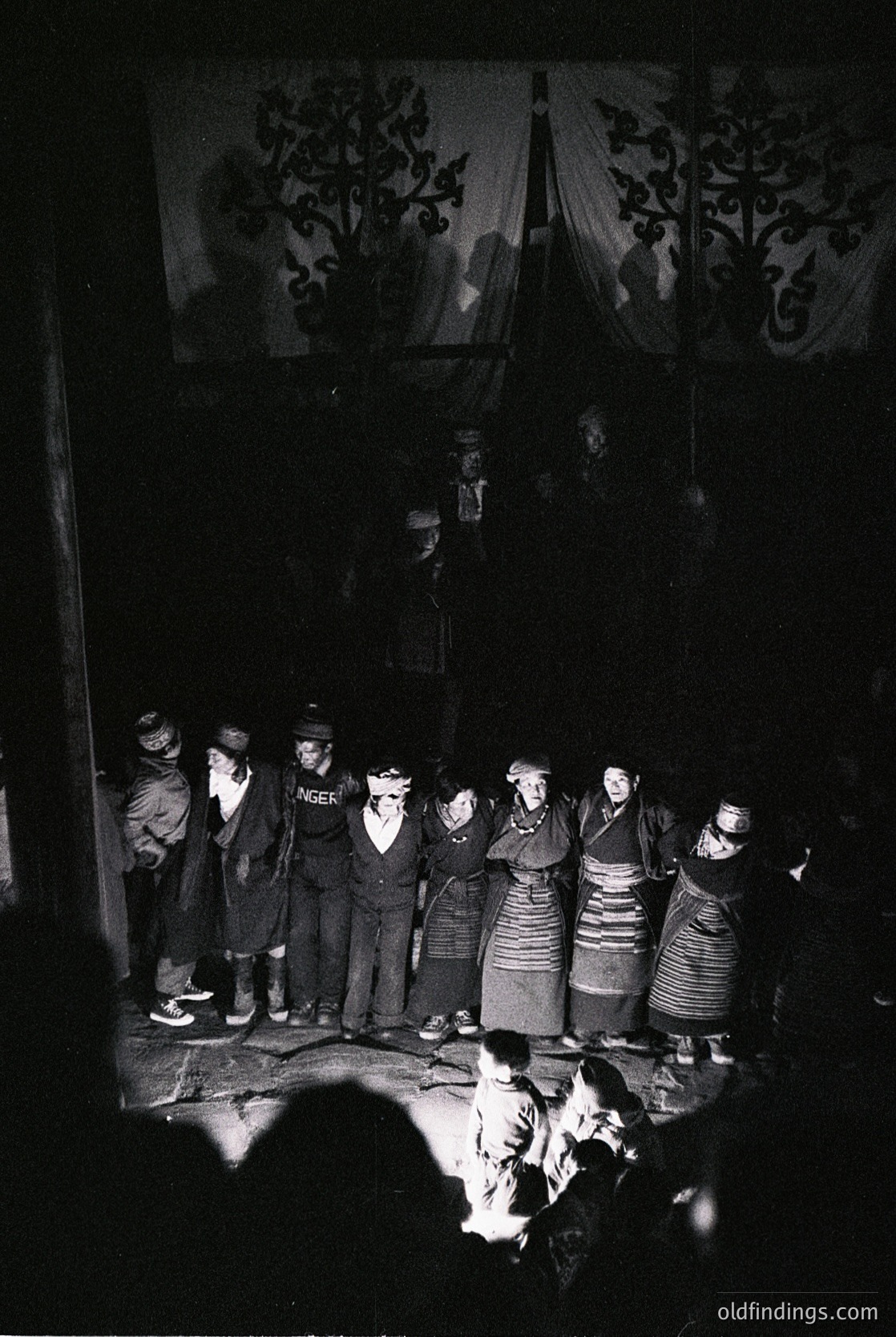 Traditional folk ensemble performing in dimly lit indoor setting, likely a rural or cultural hall. Group of 10+ individuals in matching embroidered costumes with striped aprons and headscarves, standing in a semi-circle. One central figure appears to be conducting or singing, with a banner featuring ornate floral patterns behind them. Firelight casts warm glow on faces, suggesting a communal gathering. Style and attire suggest Eastern European folk traditions, possibly or early .