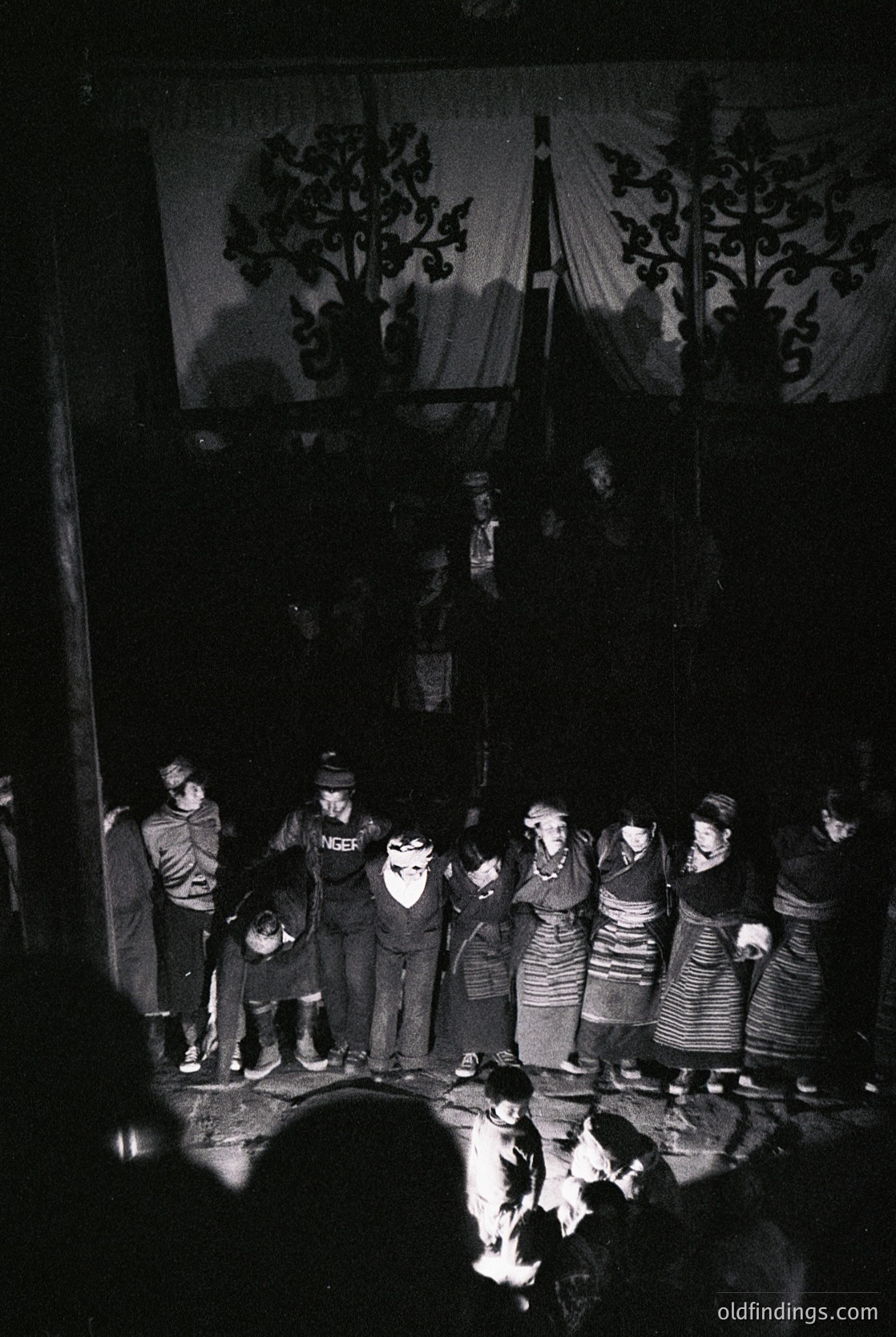 Vintage group performance in traditional folk attire, likely Eastern European (1960s–70s). Men in dark suits with hats, women in embroidered skirts and headscarves. Stage backdrop features ornate tree motifs. Firelight illuminates faces, suggesting a rural or folk festival setting.