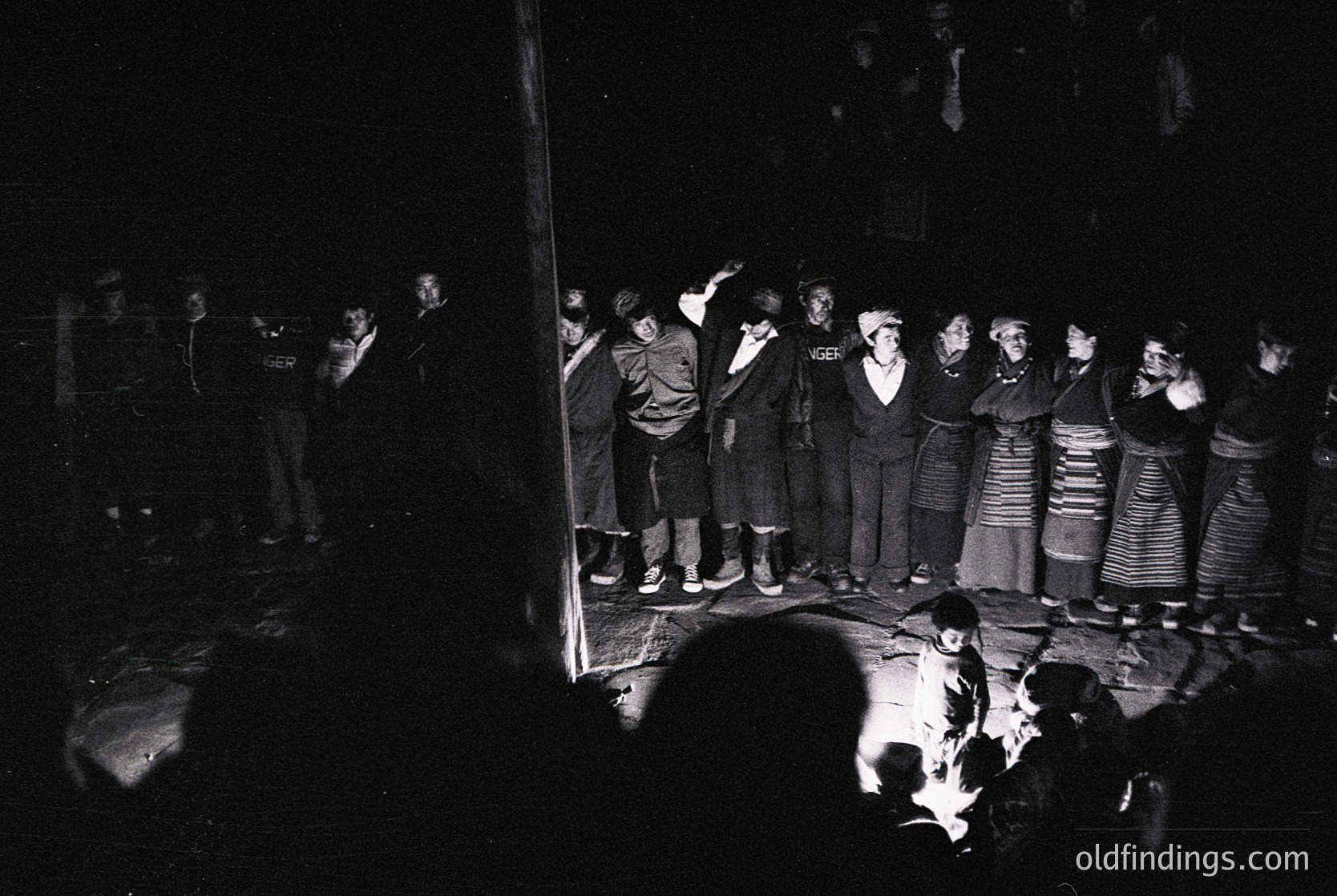 Black-and-white photo of a traditional folk choir performance in a dimly lit hall, likely Eastern European. Women in striped folk dresses and men in dark suits stand in a semi-circle, singing or clapping. A central figure plays a stringed instrument, while an audience sits in shadowed rows. Style suggests 1960s–1980s cultural preservation era.