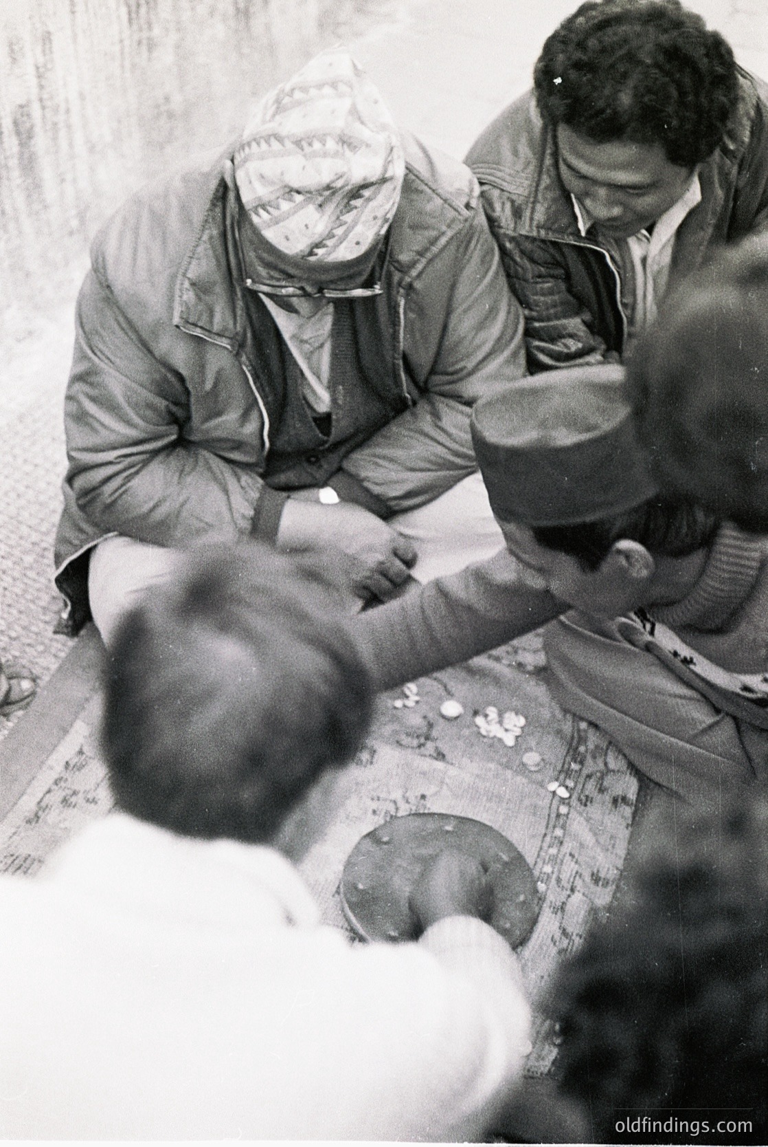 Black-and-white street scene from the 1960s–70s, likely Eastern Europe. Three men in winter coats and headwear gather around a table with coins, suggesting a street vendor or informal transaction. Urban setting with rough pavement and indistinct buildings in background.