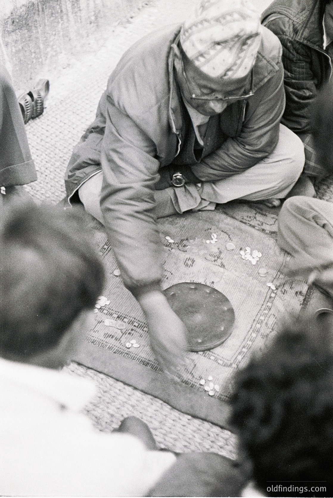 Mid-20th century black-and-white photo of a man in a white cap and glasses meticulously sorting small metal discs on a patterned cloth. Likely a factory or workshop setting, suggesting precision work.