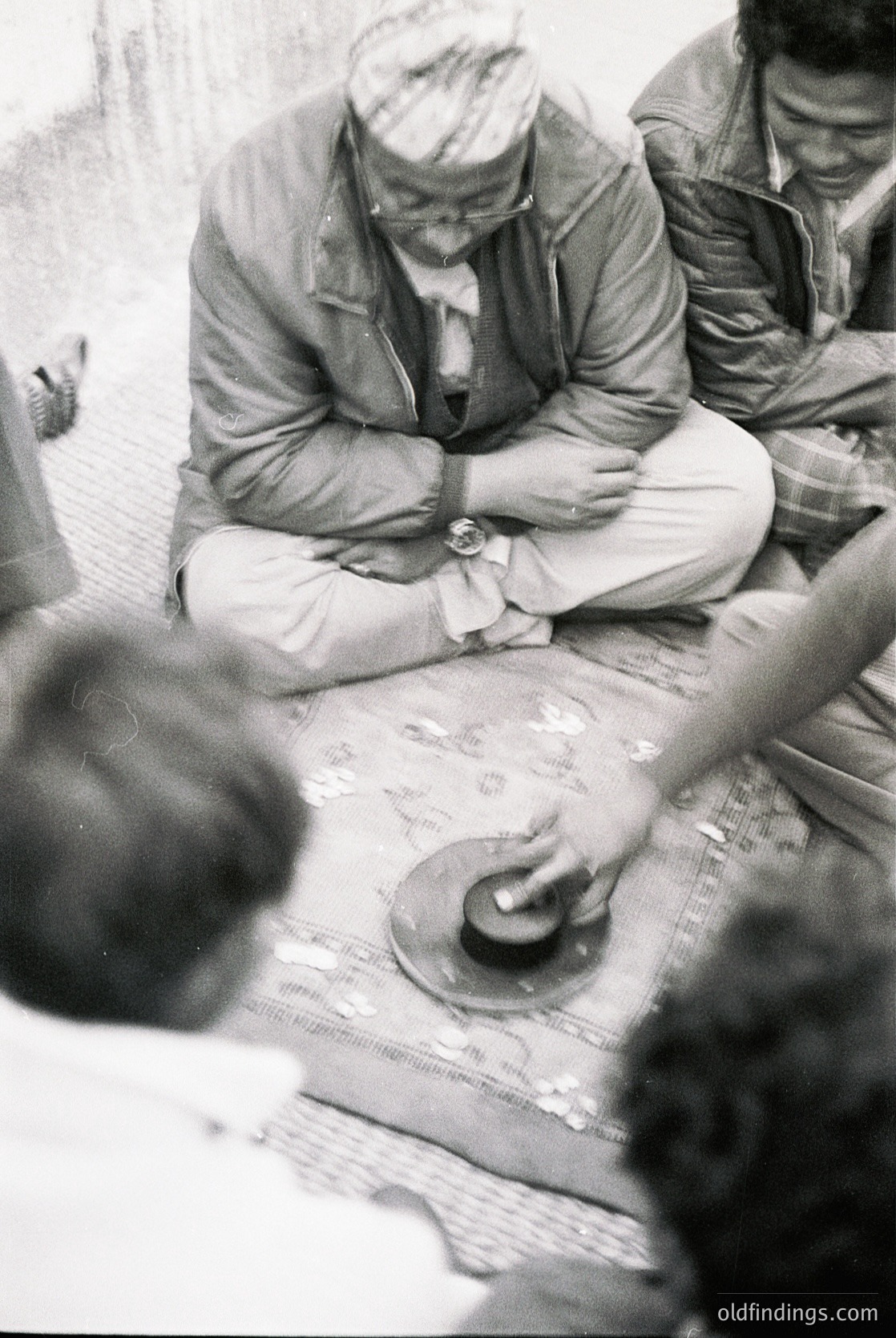 Men seated around a large, folded map in an outdoor setting, likely mid-20th century. Foreground shows a watch and hands adjusting the map. Casual, utilitarian attire suggests military or surveying activity.