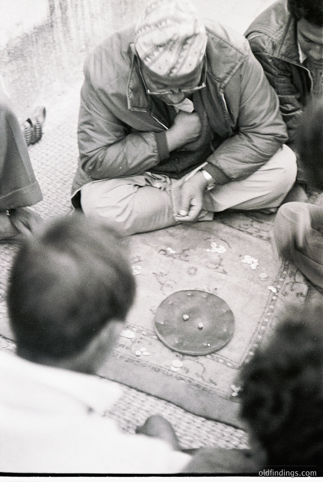 A man in 1960s-era military uniform repairs a shoe using a cobbler’s tool on a patterned floor. Blurred figures in the background suggest a public or communal space.