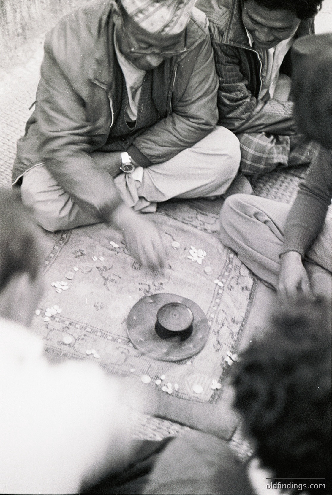 Black-and-white photo of three men playing a traditional board game on a woven mat, likely in a rural setting. Central figure wears a watch and light-colored pants, seated cross-legged. Game board features intricate designs and a central circular piece. Mid-20th century attire suggests or .